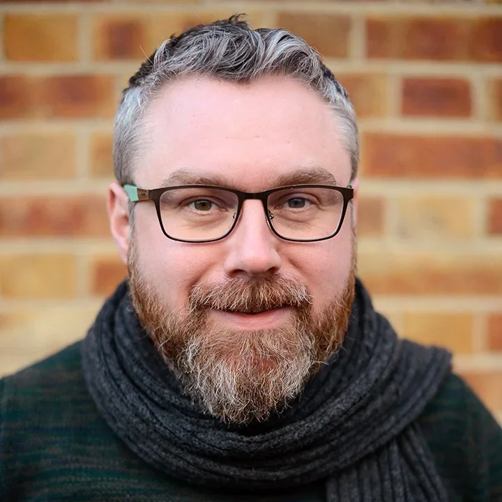 A man with glasses, a beard, and short gray hair smiling in front of a brick wall.
