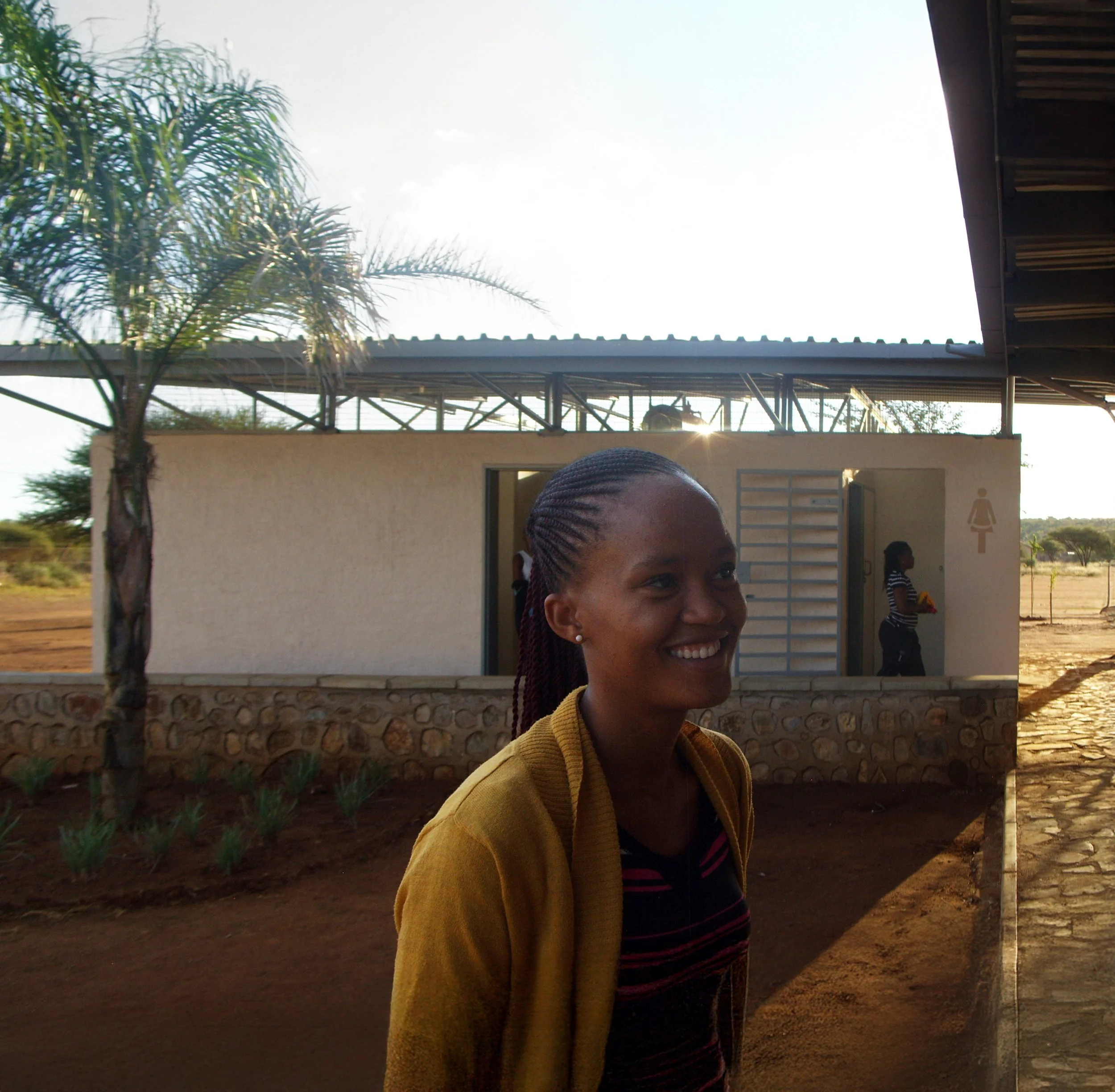 Smiling woman outdoors near bathroom with female figure in the background, palm trees, and a clear sky.