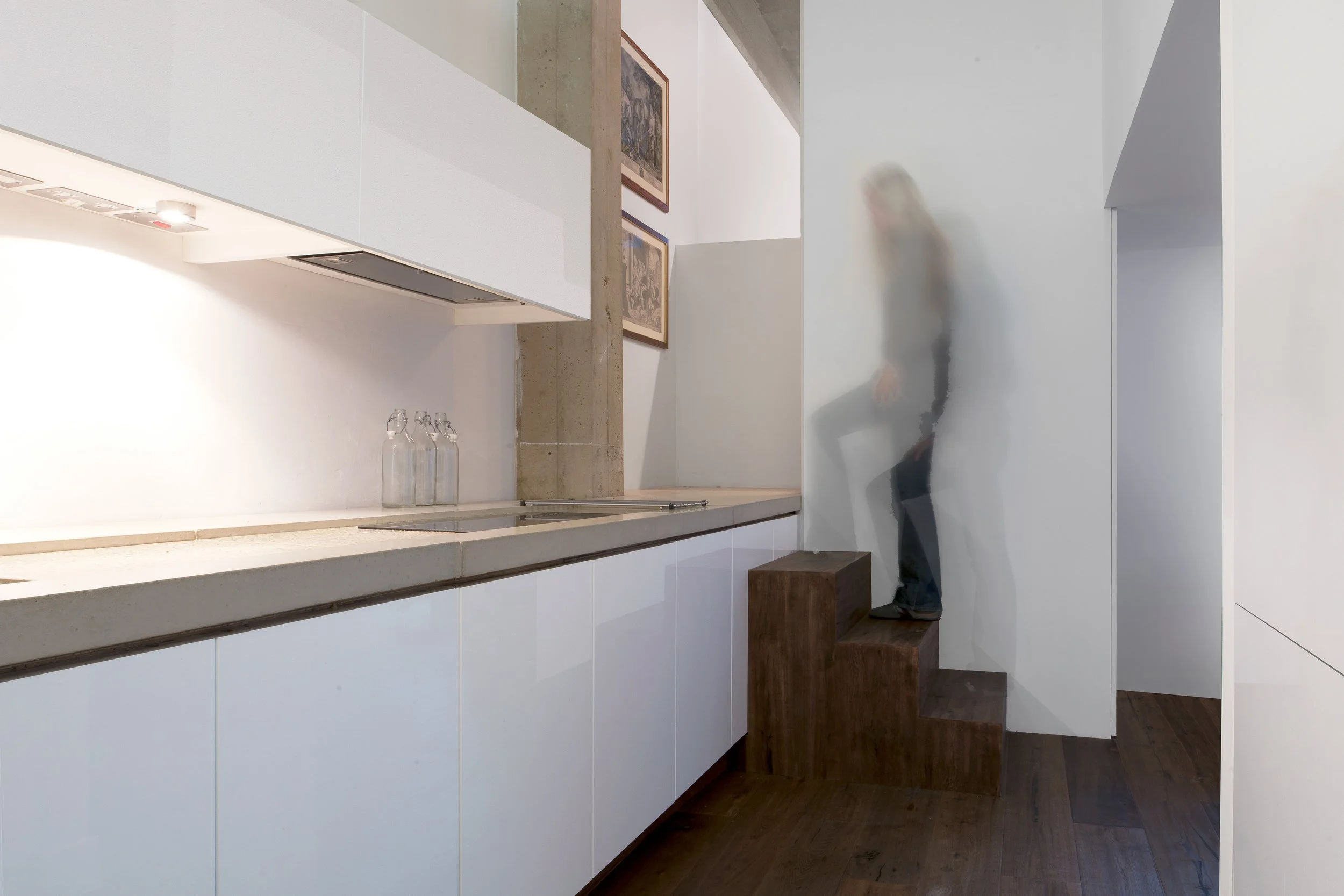A person climbing a small wooden staircase in a minimalist kitchen, with frosted glass walls and white cabinets.