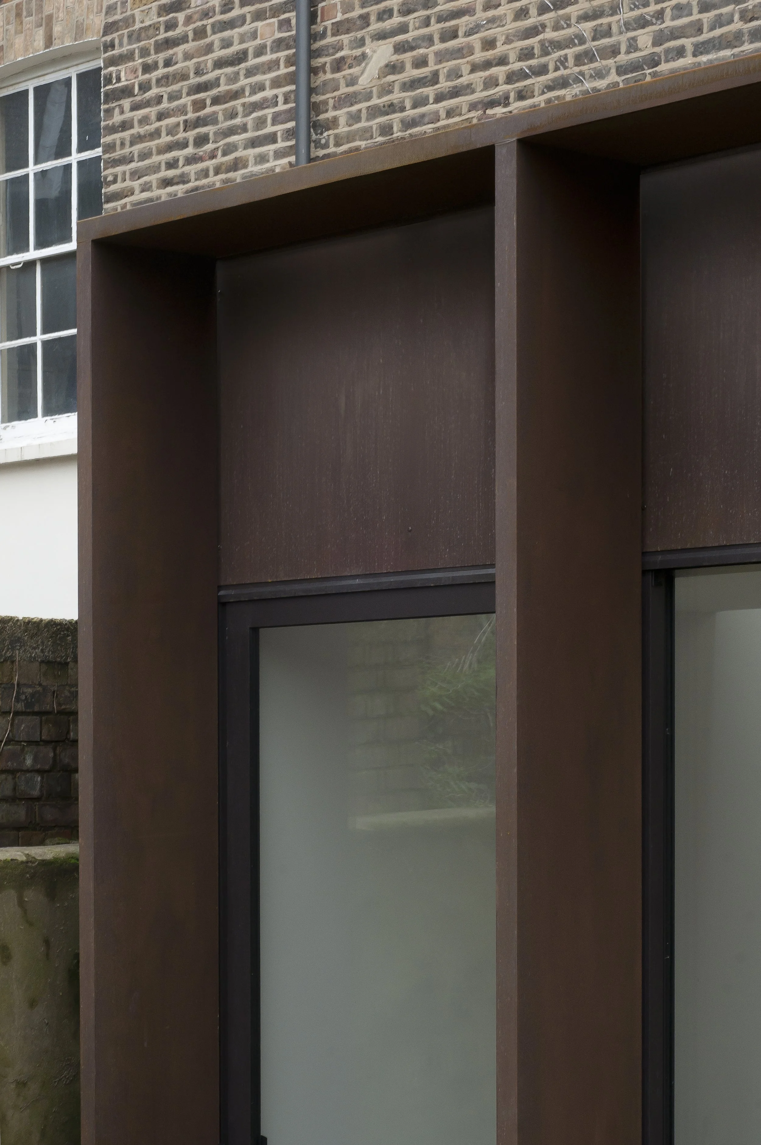 Close-up of a modern building facade with a large frosted glass window and rust-colored metal framing against a brick wall background.