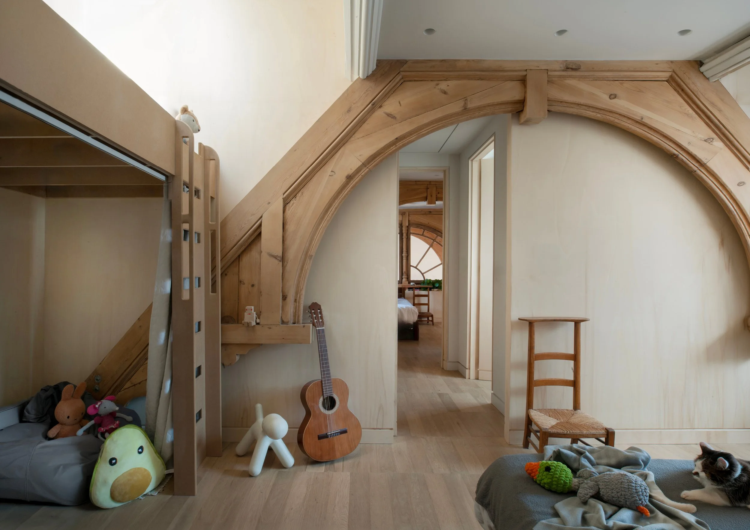 A cozy room with wooden accents and furniture, including a small bed under a bunk bed, a guitar, and a chair. Toys and plush animals are on the beds and floor, with a cat sleeping on a blanket.