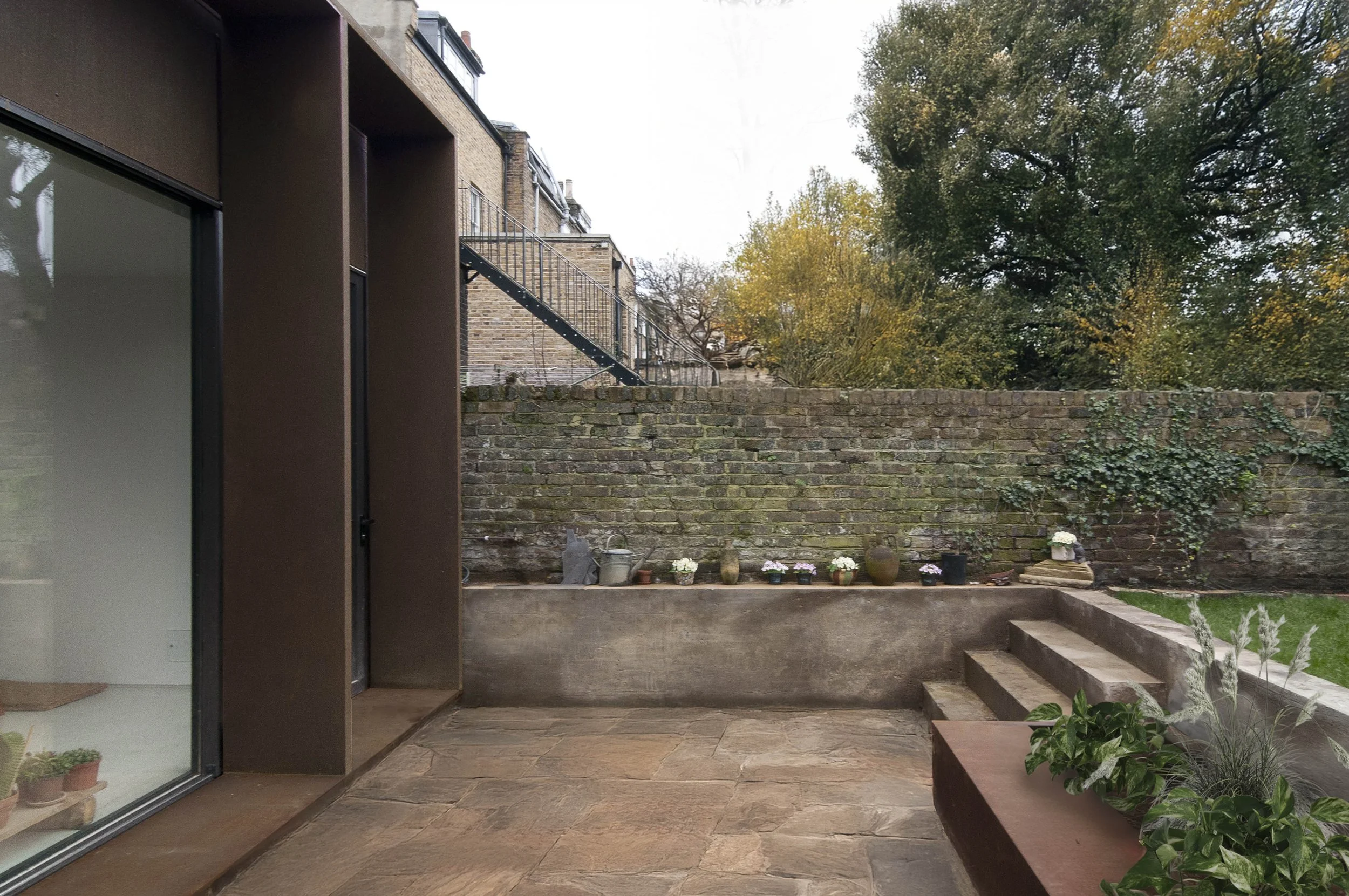 A backyard patio with stone flooring, potted plants, and a brick wall with outdoor stairs. The background features trees with fall foliage.