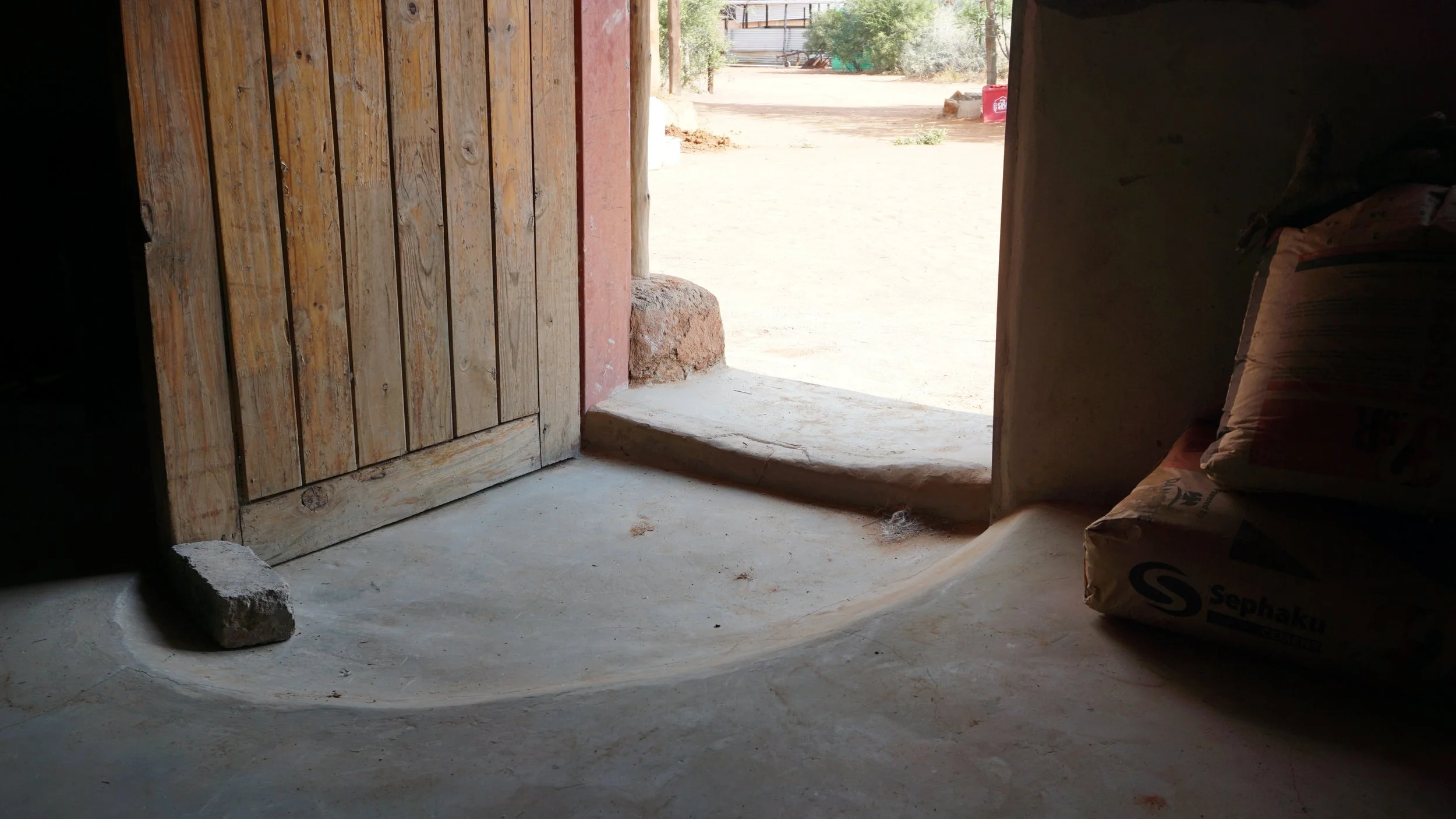 Entrance to a room with a wooden door, estimate of sunlight coming in, concrete floor, and a stack of cement bags on the right side.