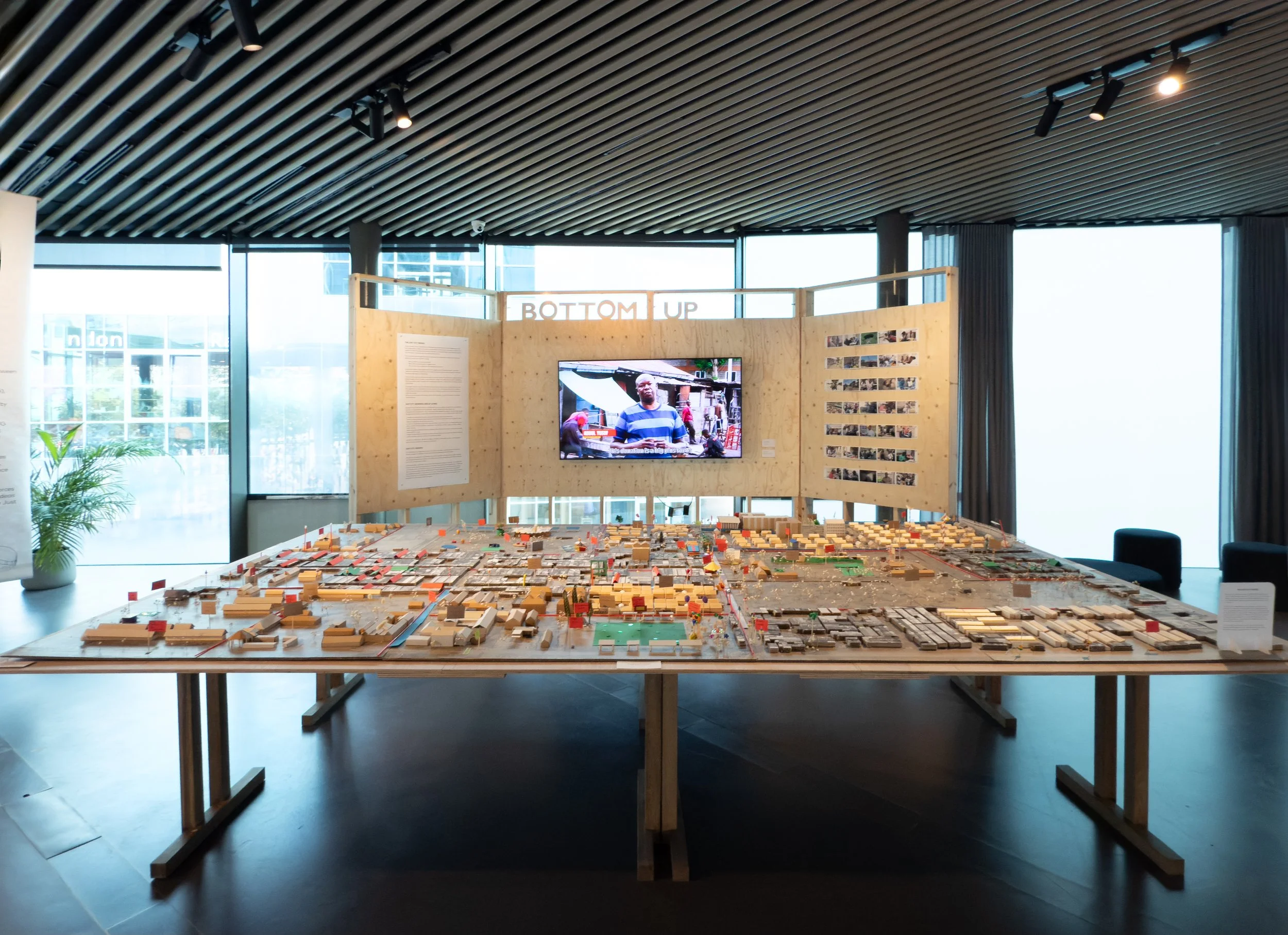 A large detailed architectural model displayed on a wooden table inside a modern building with floor-to-ceiling windows, black ceiling, and wooden slat ceiling, featuring a large video screen and informational panels in the background.