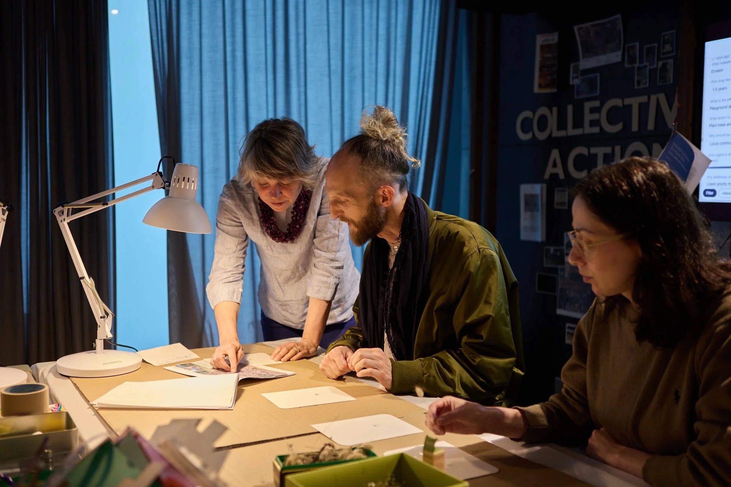 Three people working together at a table covered with papers and documents in a dimly lit room with blue curtains. One woman is standing and pointing at something on the table, while a man and another woman are seated and looking at the papers.