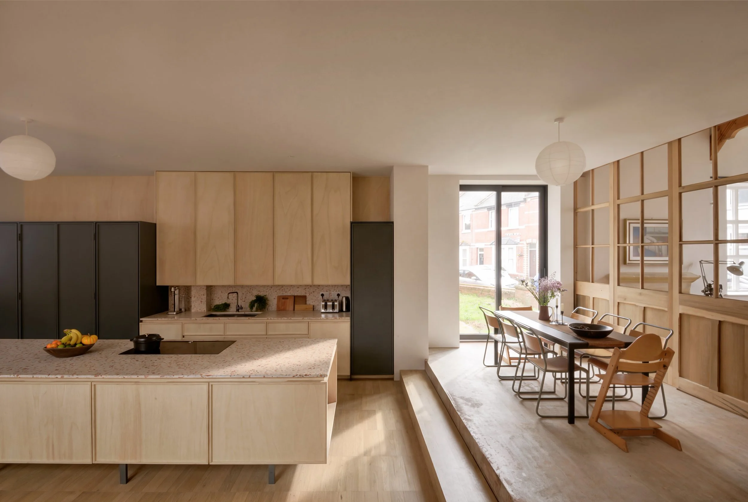 Modern kitchen and dining area with wooden cabinets, a breakfast bar with fruit, and a dining table with chairs near large windows allowing natural light.