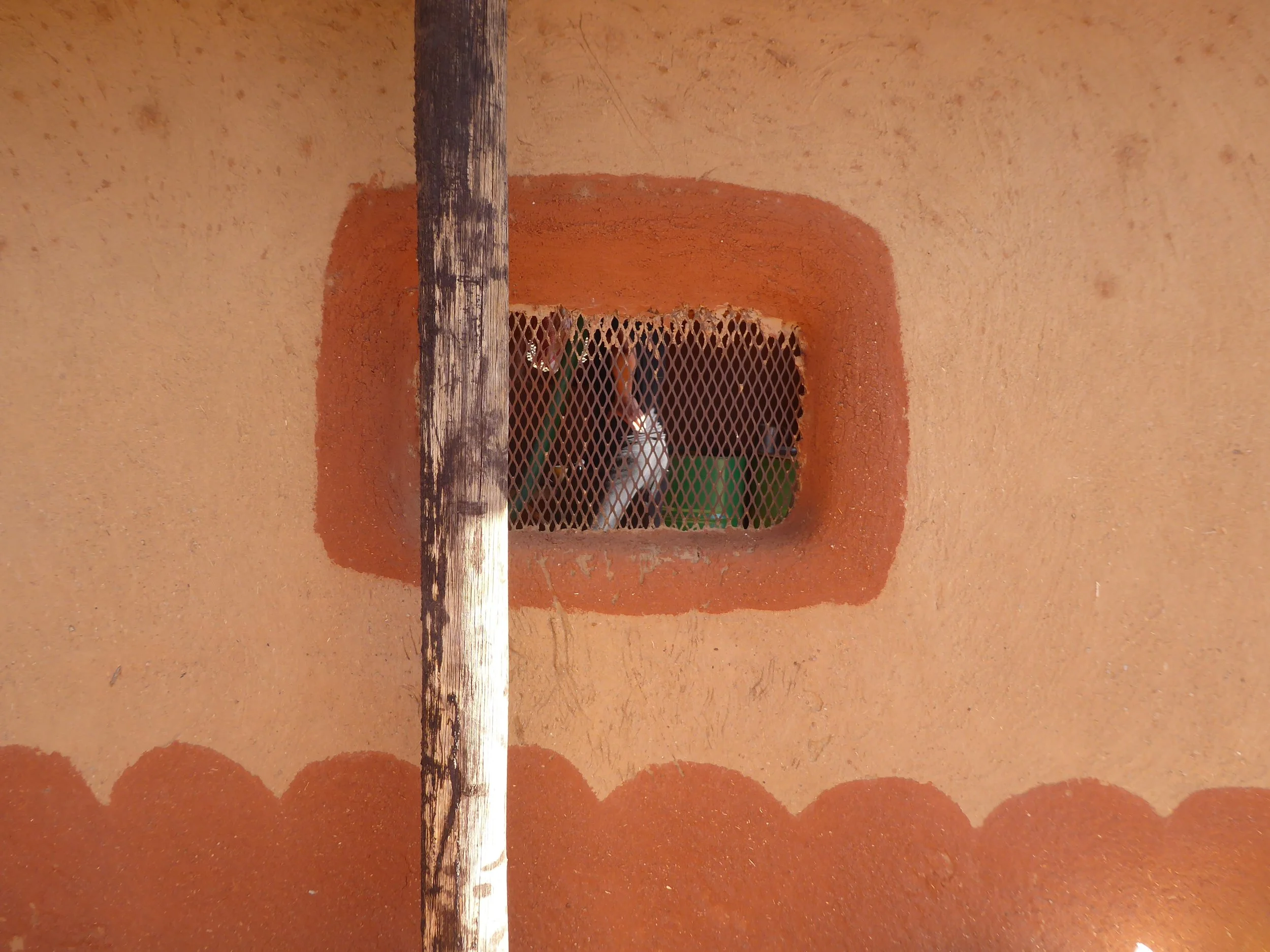 View looking up at a small, rectangular opening with a metal grate. Behind the grate, a white cat is peeking through, and part of a green object is visible inside.