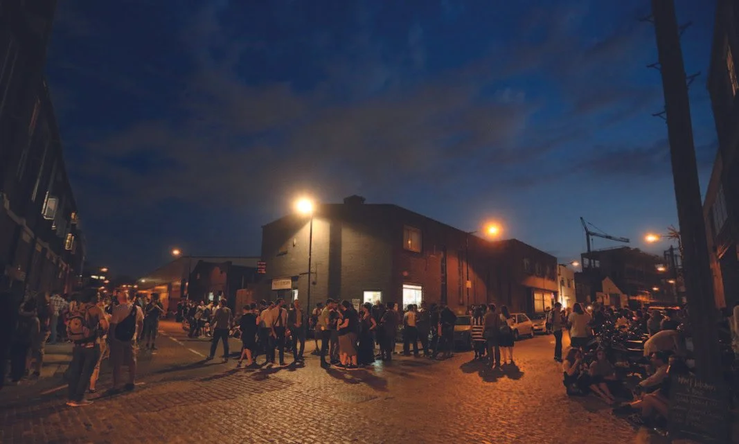 Nighttime scene of a busy street with a crowd of people gathered, illuminated by streetlights, against a dark sky with clouds, and buildings on both sides.