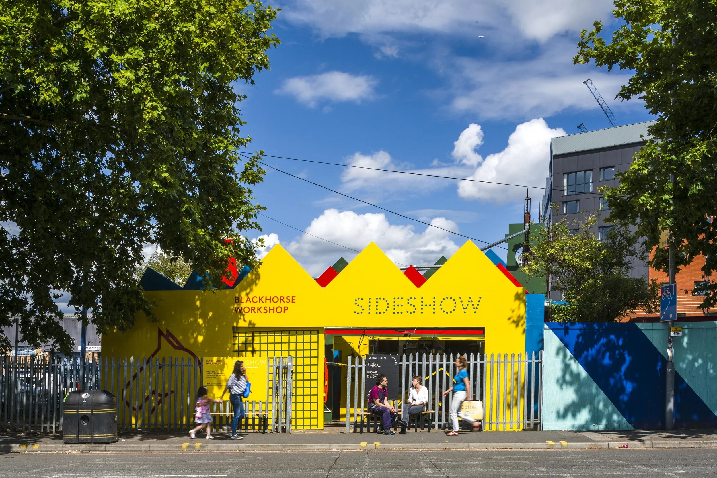 A colorful urban scene featuring a bright yellow building labeled 'SIDESHOW' with palm-shaped peaks, surrounded by trees and modern buildings under a partly cloudy sky. There are people sitting and walking outside the building, and a fence with a small black chalkboard sign.
