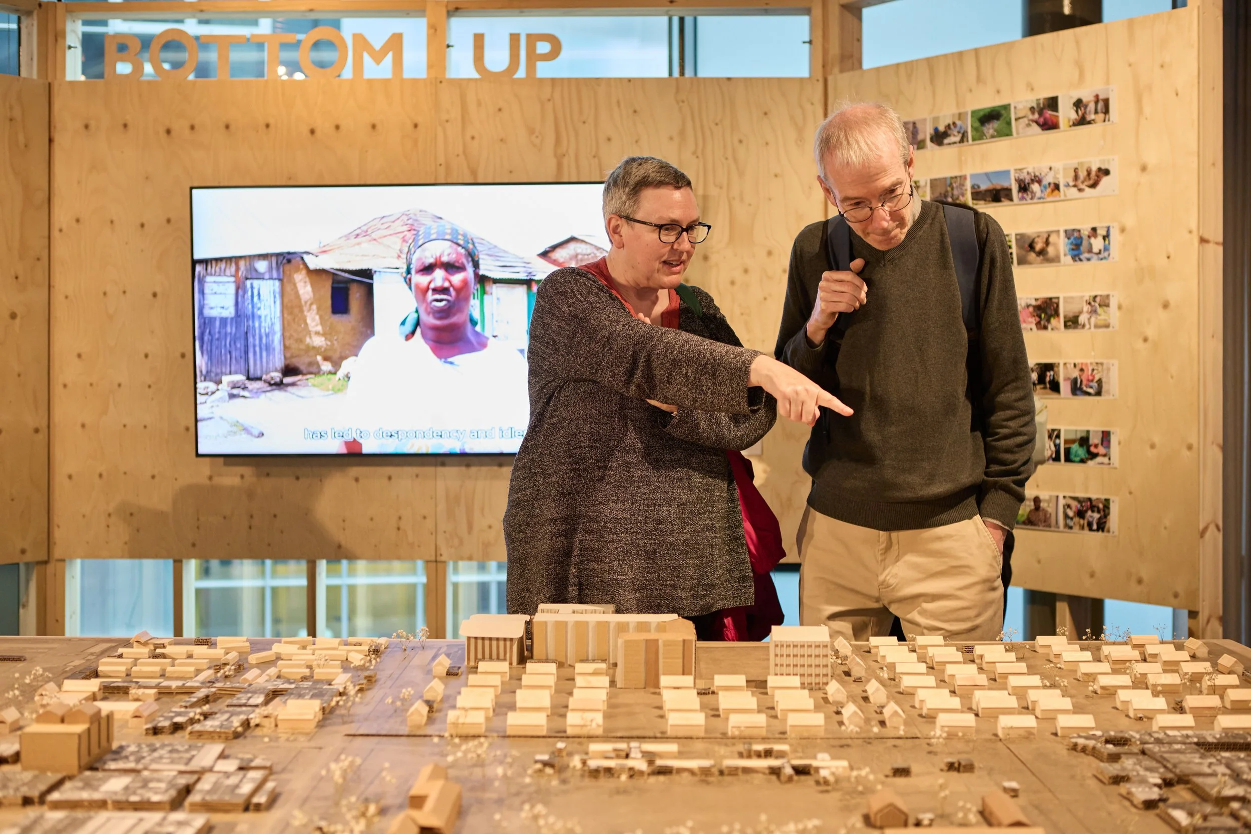 Two people, a woman and a man, are standing and looking at a detailed architectural model of a city or neighborhood on a table. The woman is pointing at the model, and is wearing glasses and a dark coat. The man is carrying a backpack, wearing glasses and beige pants. Behind them, there is a large screen displaying a photo of a woman with an older, wooden building in the background, and some smaller photos are pinned on a board to the right.