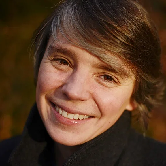 A young man with short brown hair smiling outdoors during sunset, wearing a black sweater.