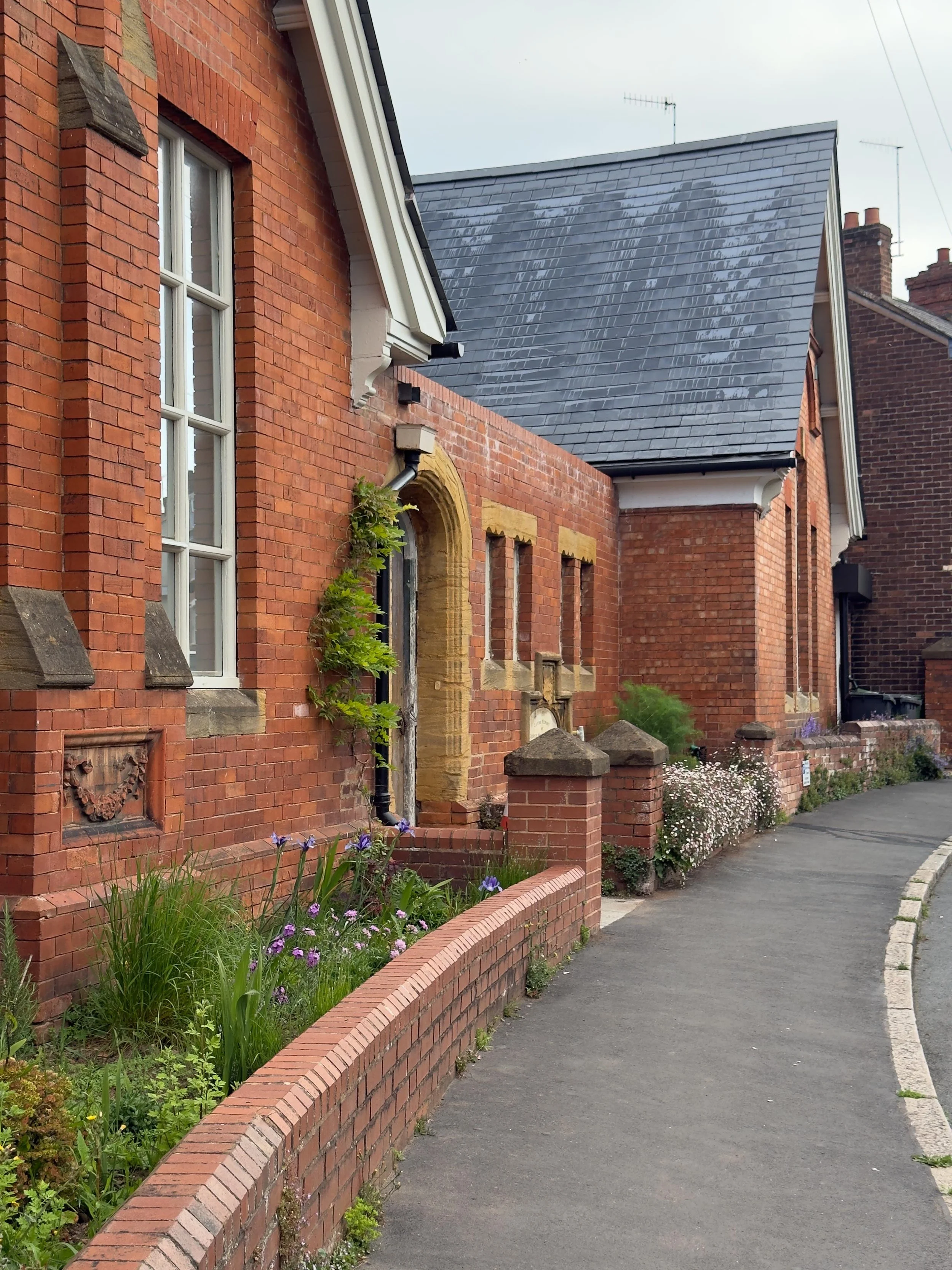 A brick house with tall white window panes, green plants, and flowers along a curved sidewalk.