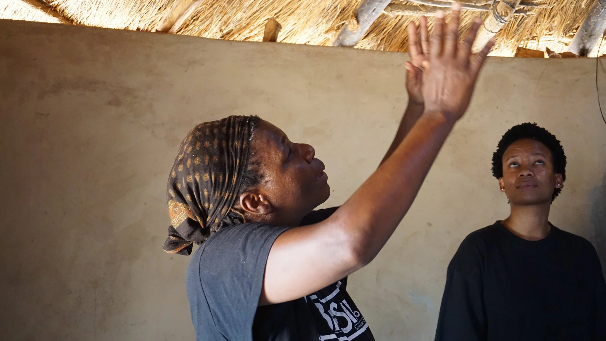Two women inside a room with a thatched roof, one woman is raising her hand towards the ceiling while the other woman stands nearby looking up.