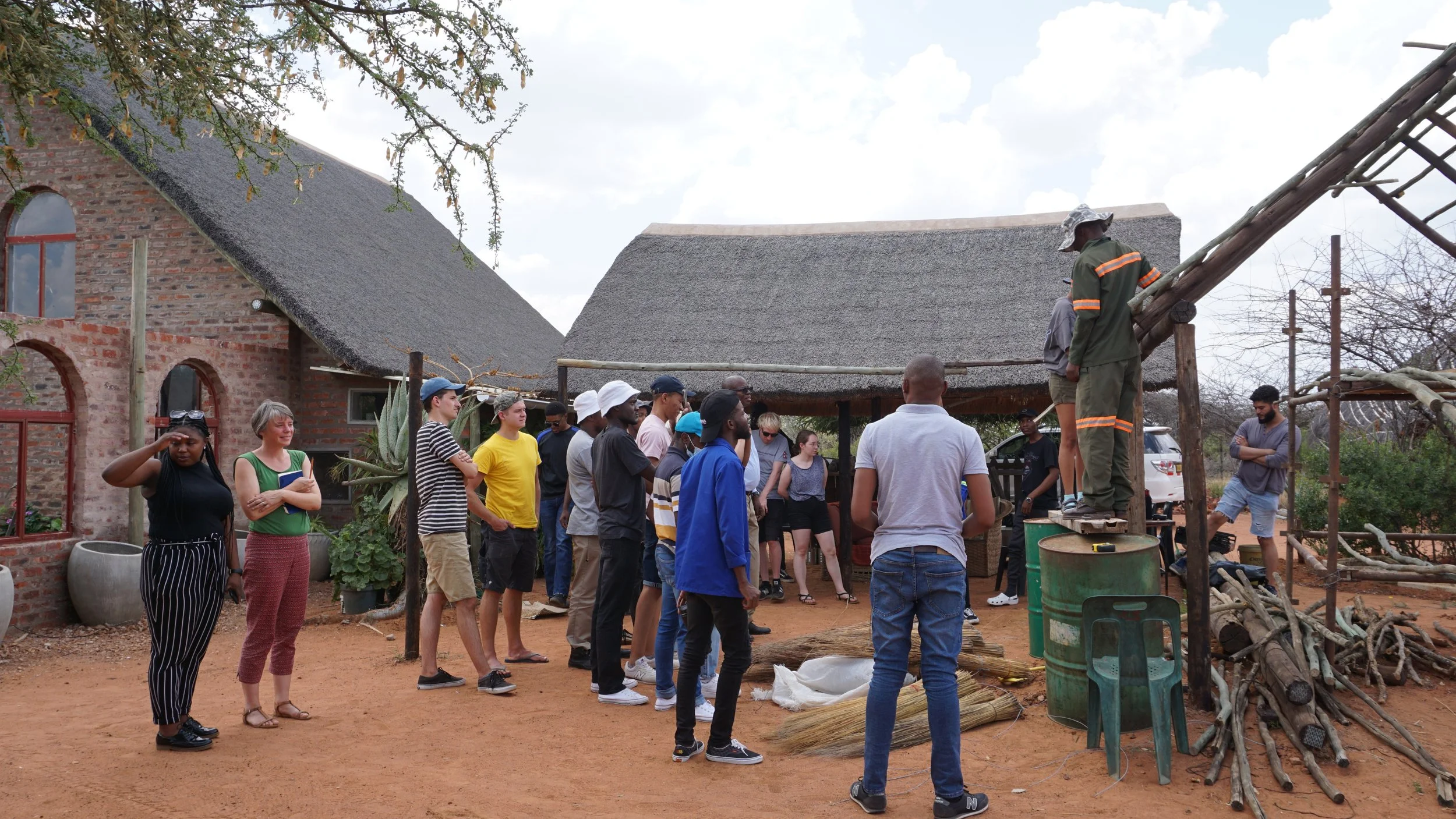 Group of people gathered outside a building with thatched roofs, listening to a speaker standing on a wooden platform, with a firefighter in uniform helping.