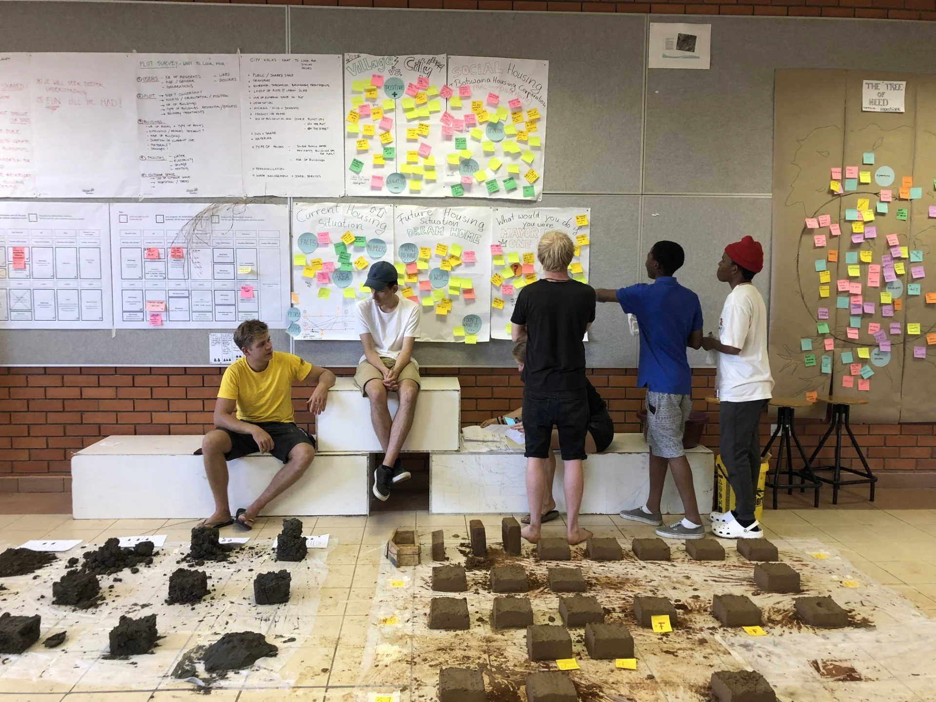 Group of young people participating in a community planning event. Some are sitting on white tiered blocks, while others are standing and discussing. The background features large sheets of paper and posters with colorful sticky notes and handwritten