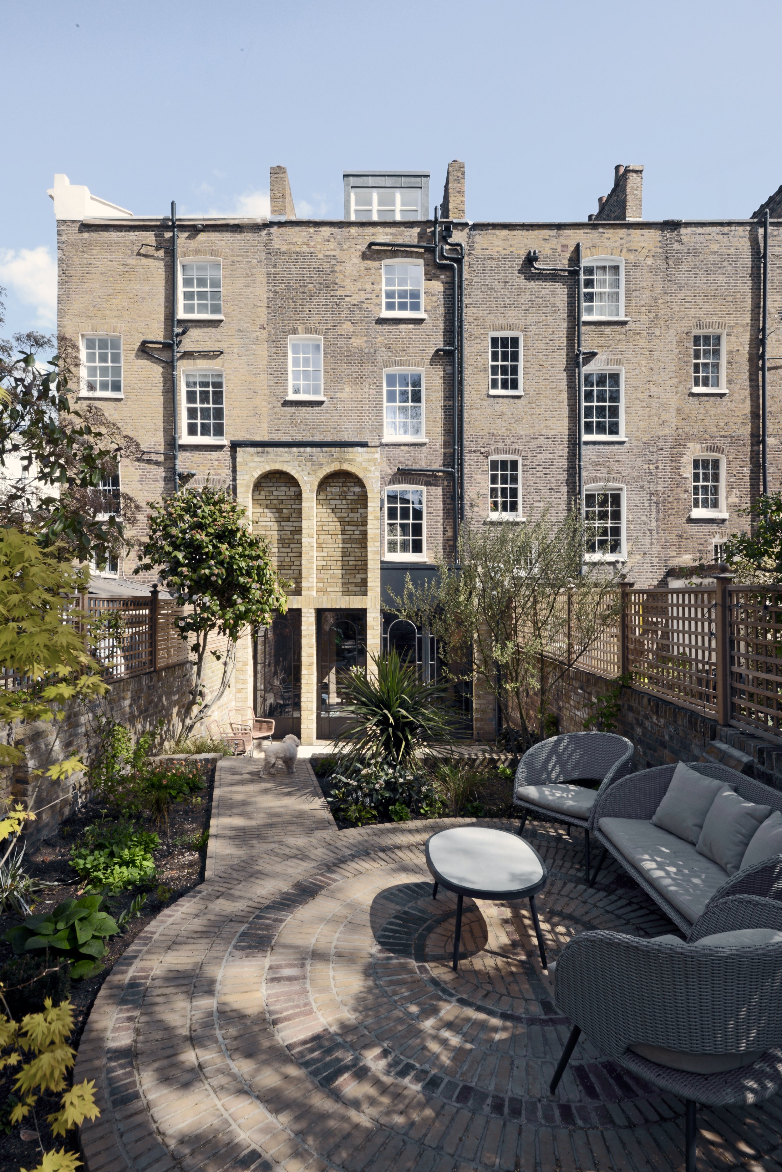 Backyard patio with rattan furniture, brick pathway, garden plants, and a small white dog, with a multi-story brick apartment building in the background.