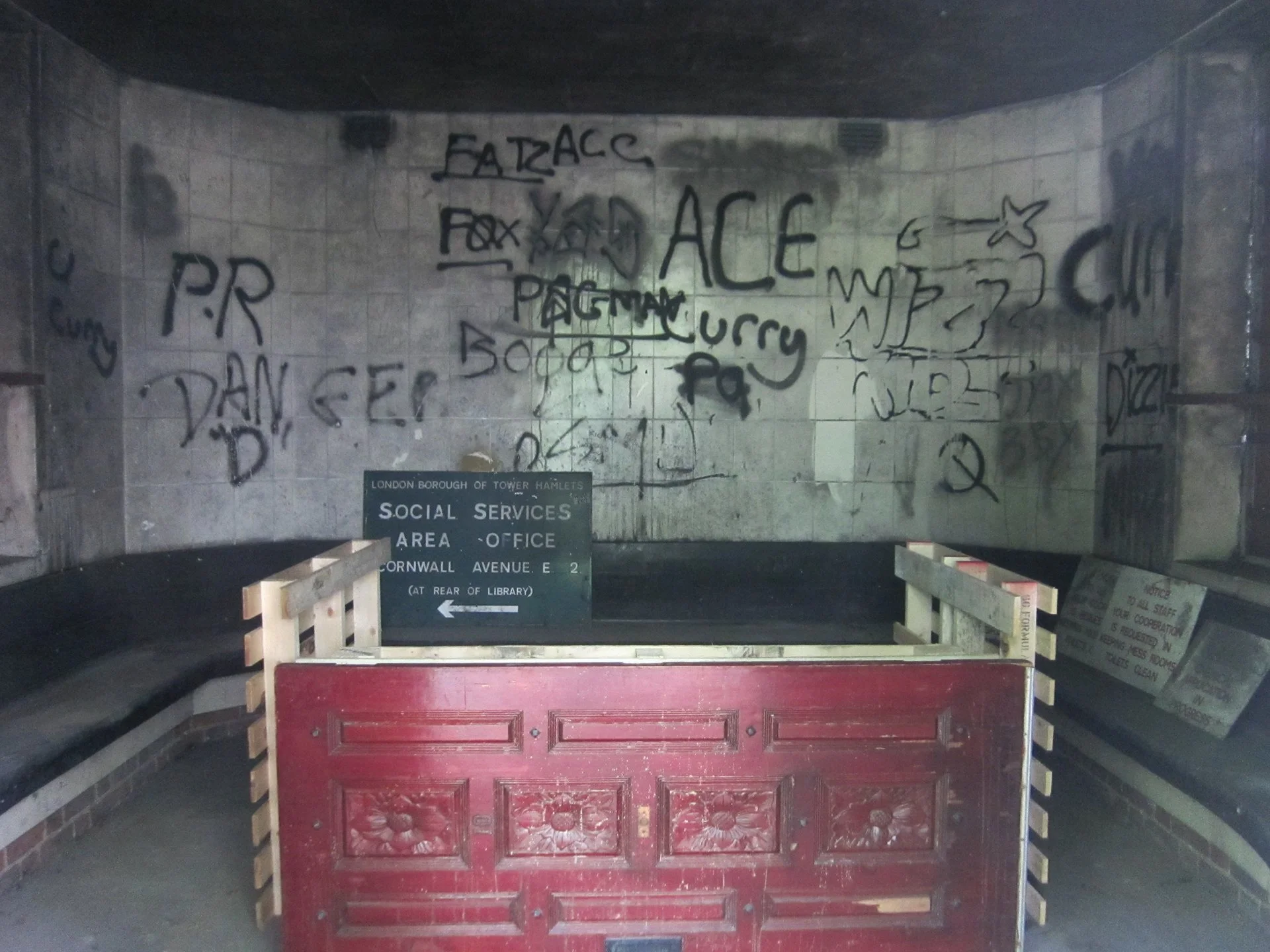 A subway entrance with graffiti-covered walls and a sign for the London Borough of Tower Hamlets Social Services Area Office on Cornwall Avenue E2, with a partially built red barrier in front.