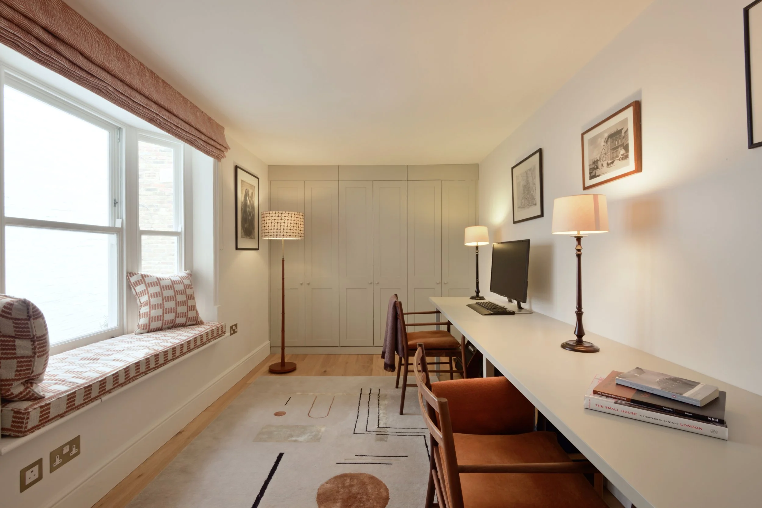 A cozy home office with a window seat, a desk with a computer, and framed artwork on the wall.