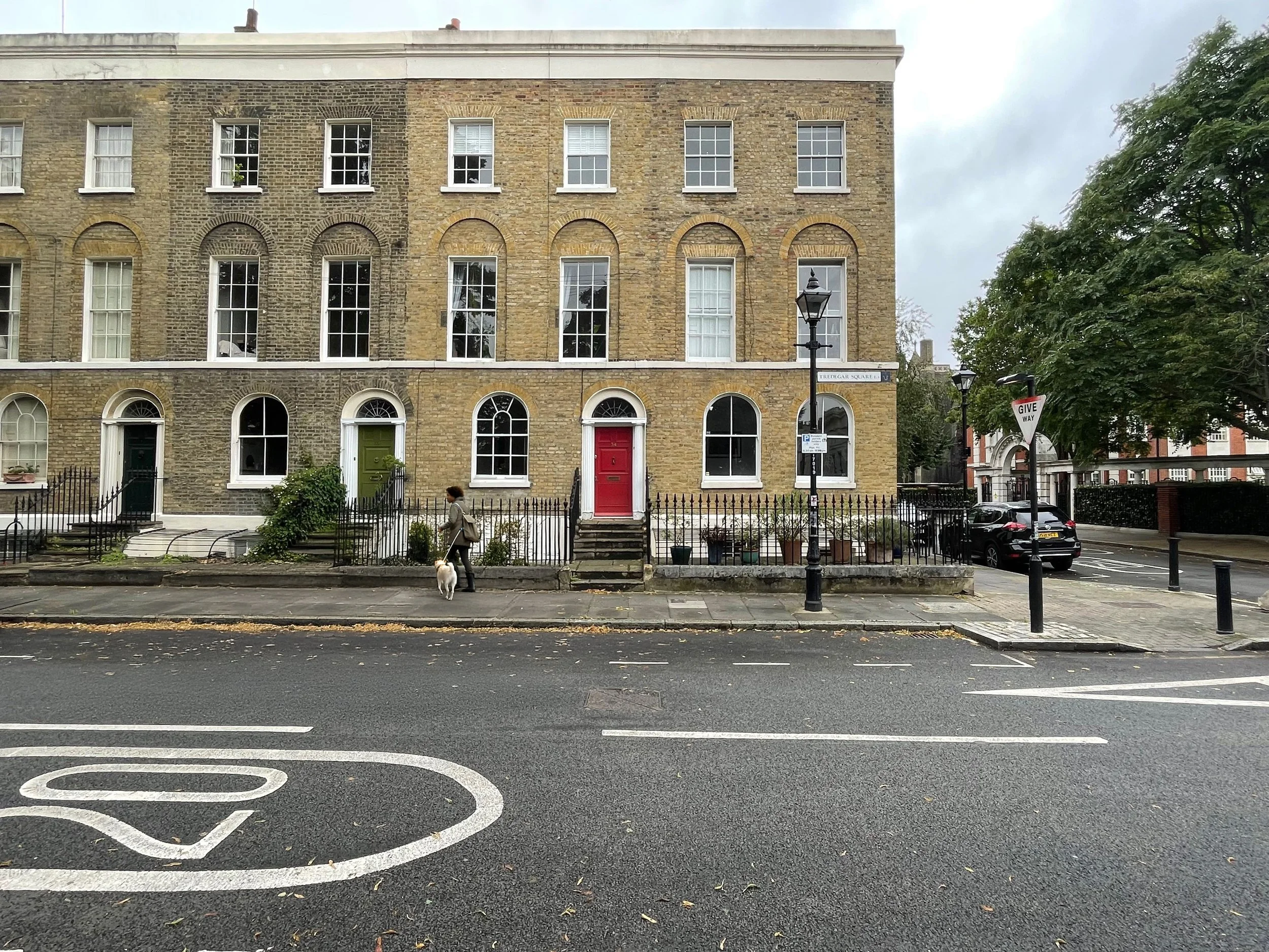A woman walking her dog on the sidewalk in front of a multi-story brick building with various colored doors and large windows. There are street signs, trees, and parked cars nearby on a cloudy day.