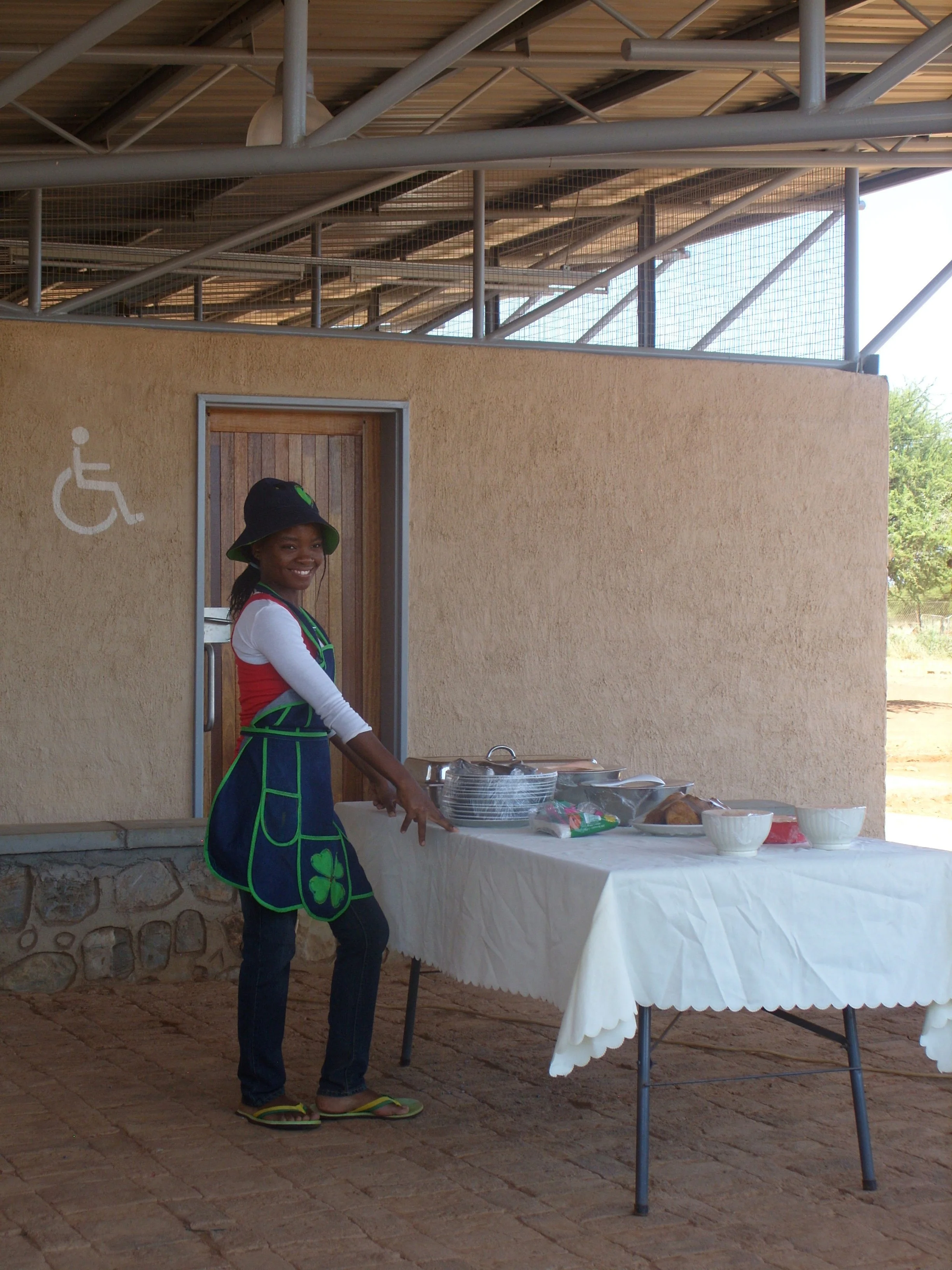 A young girl smiling and standing next to a table with food and utensils outdoors, near a building with a wheelchair accessible sign.