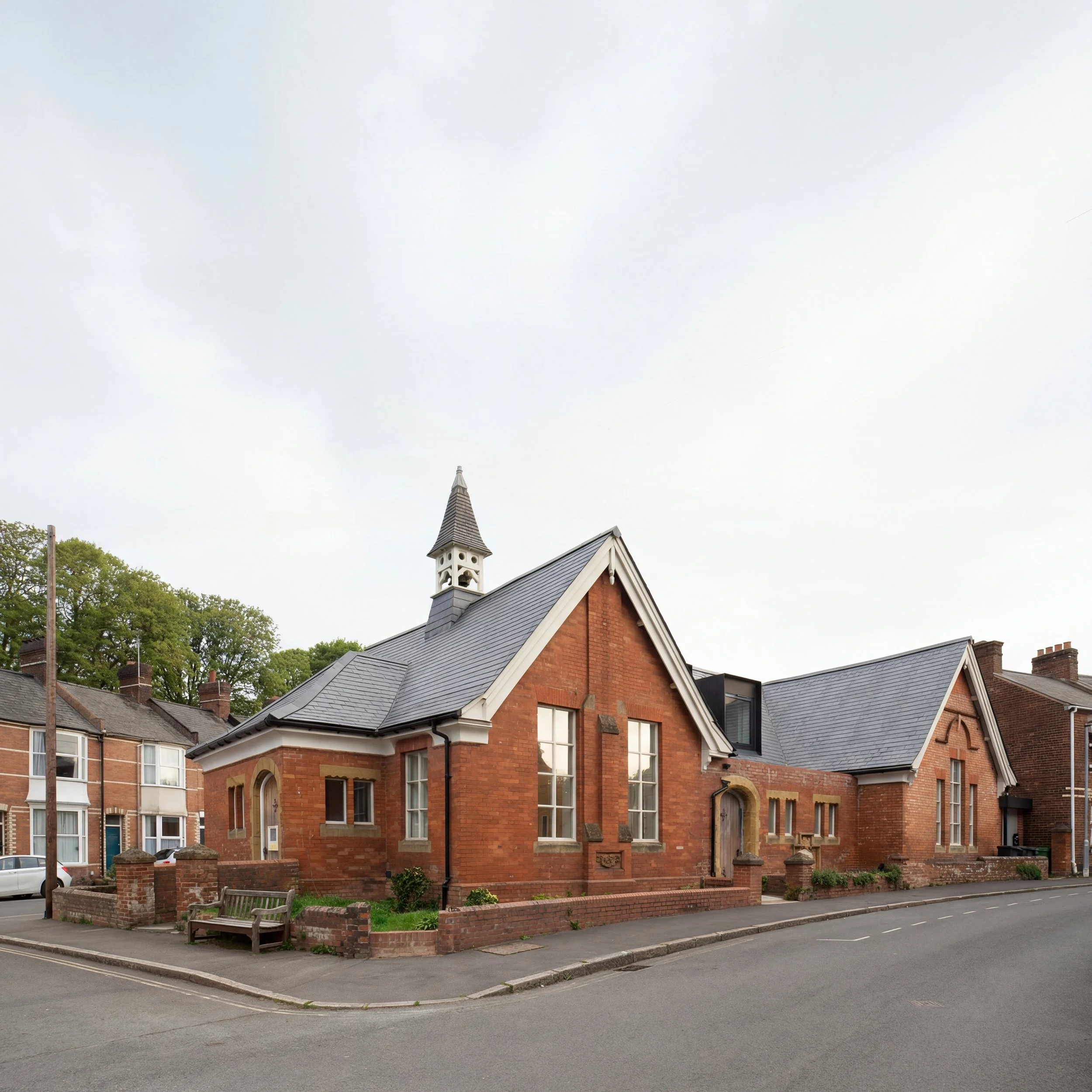 A red brick church with white trim and a tall steeple, situated along a street with a small parking area and trees in the background.