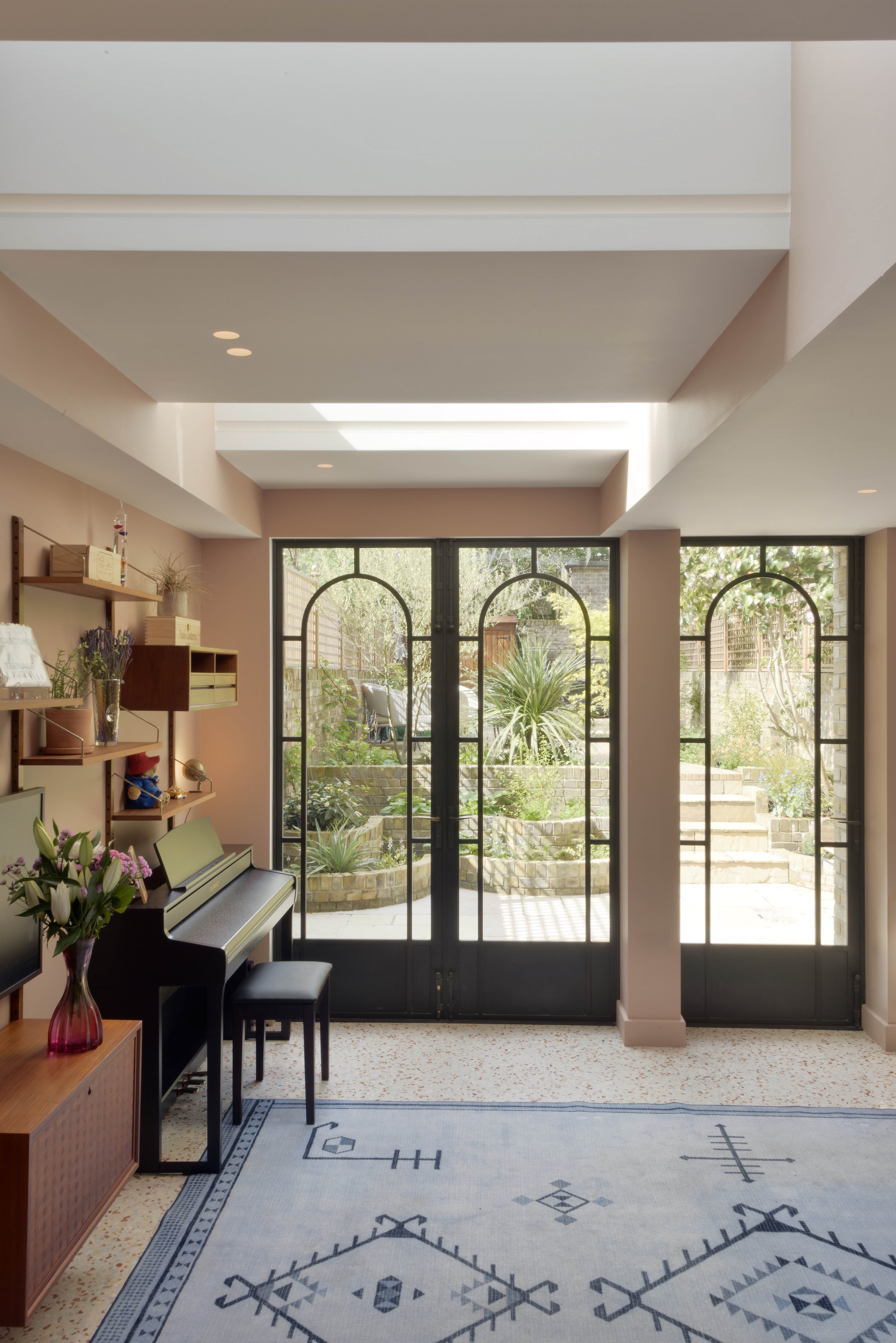 Bright living room with large glass doors opening to a garden with greenery and brick steps, featuring a black upright piano, a pink vase with flowers, and decorative shelves on the wall.