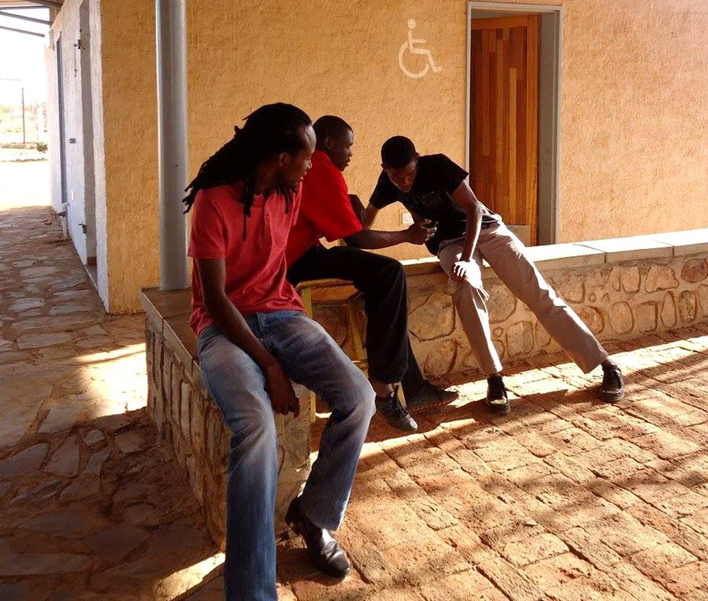 Three people sitting and leaning against a yellow wall with a wheelchair accessible sign. Two are sitting on a stone low wall, one is standing and leaning over, all looking at a phone together.