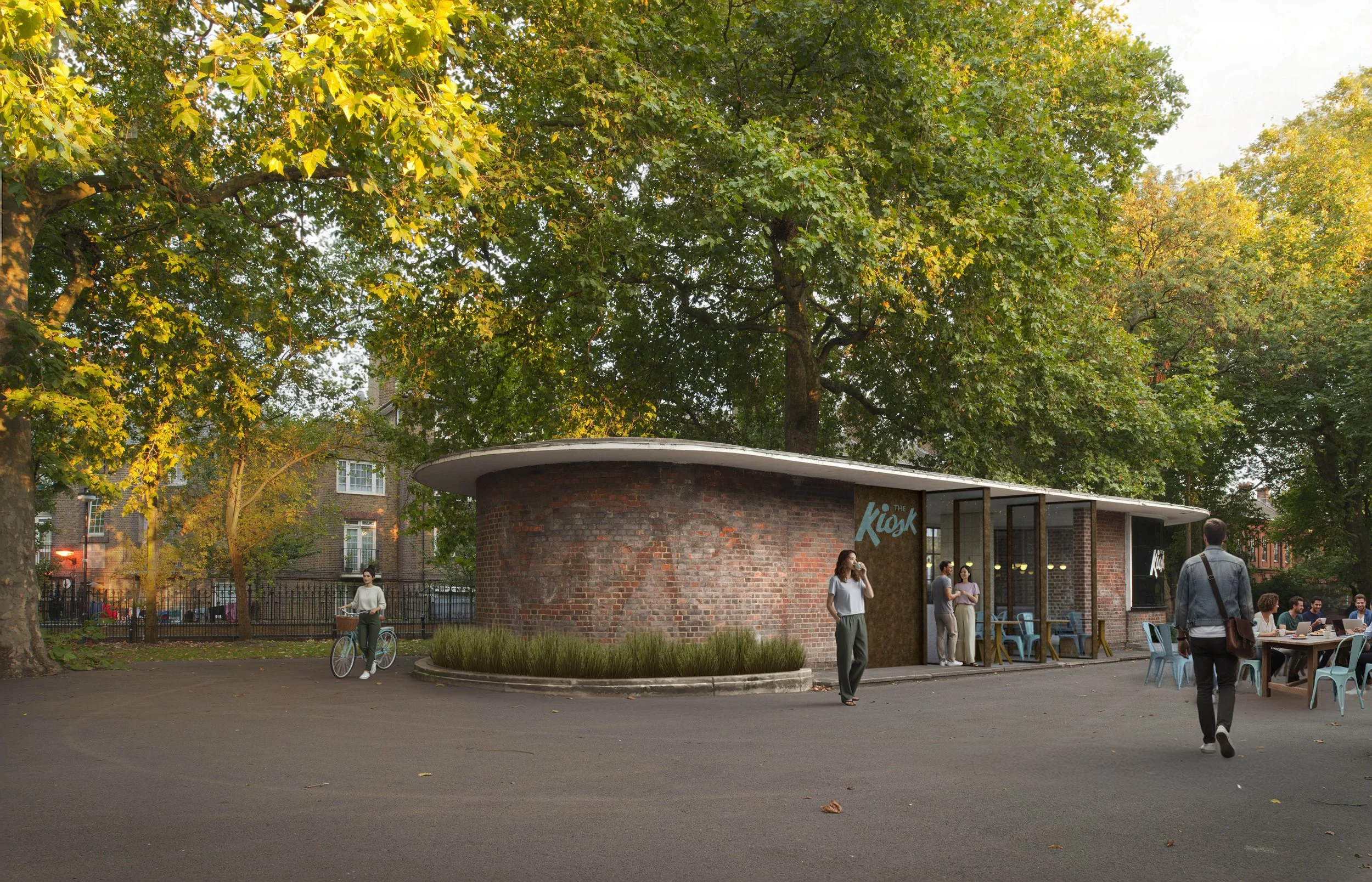 A modern brick building with a sign reading 'The Kiosk', people sitting and standing outside, one person with a bicycle, surrounded by trees with green and yellow leaves, in a park or plaza setting during autumn.
