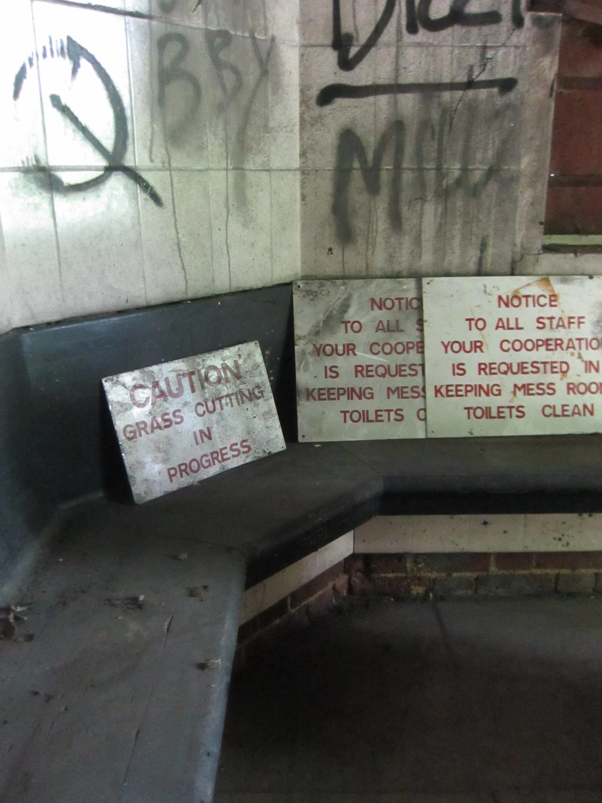A bench with three signs, one warning about grass cutting in progress, and two requesting staff cooperation to keep the mess room toilets clean. There is graffiti on the tiled wall above the bench and a clock drawing on the wall.