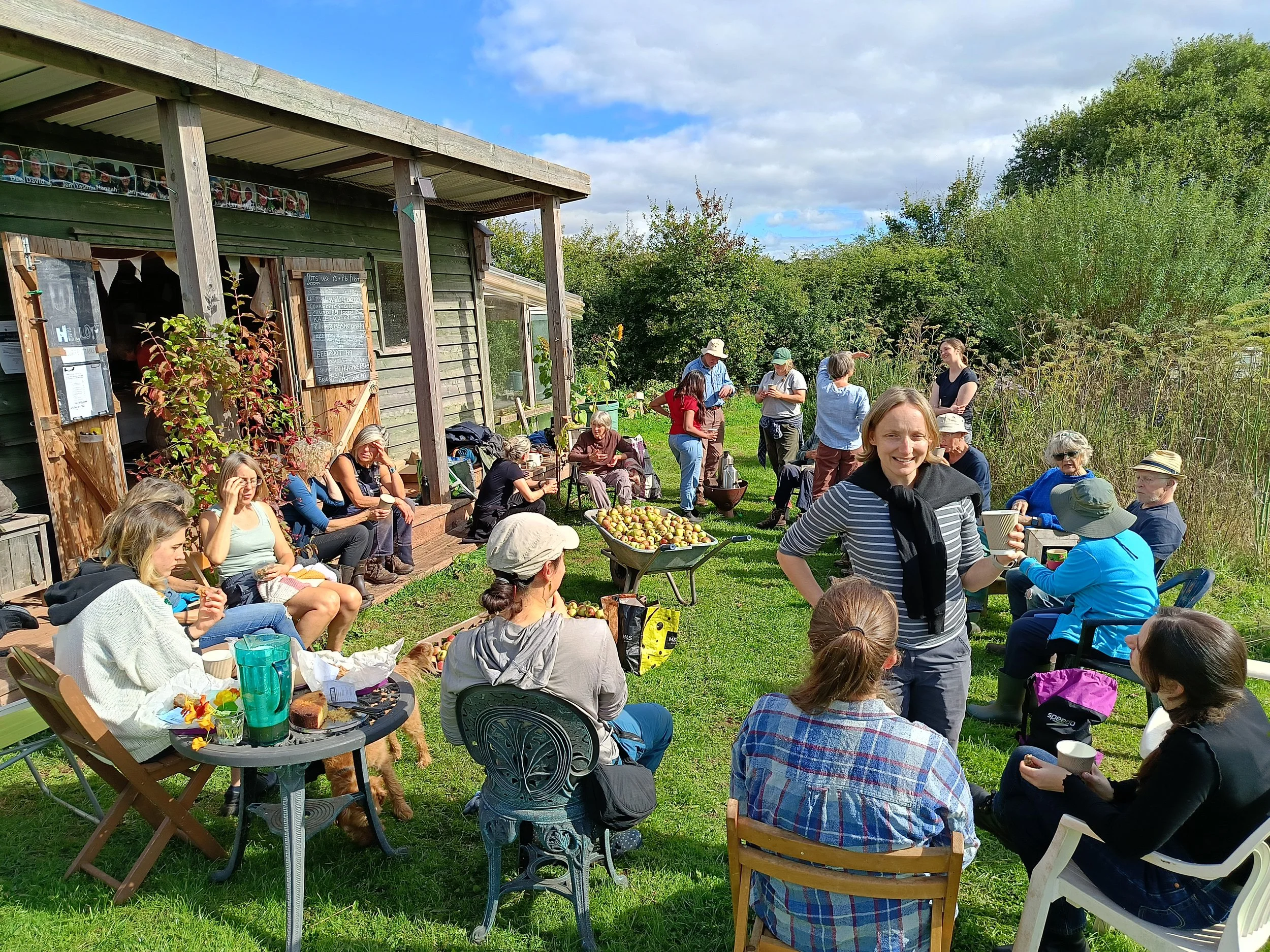 Group of people gathering outdoors on a sunny day, sitting and standing on grass near a rustic wooden building, enjoying food and drinks, with trees and bushes in the background.