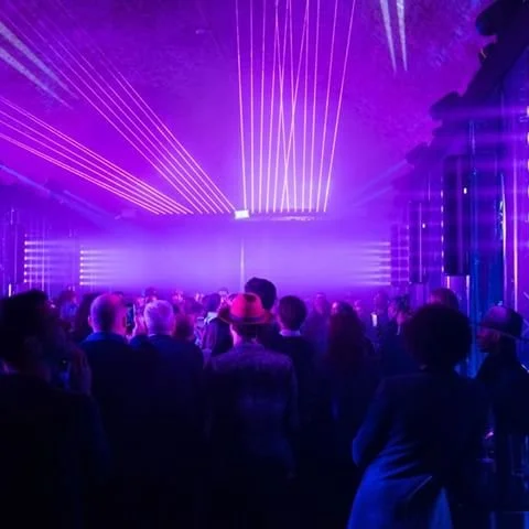 Crowd at a concert or nightclub with purple laser lights and stage lighting.