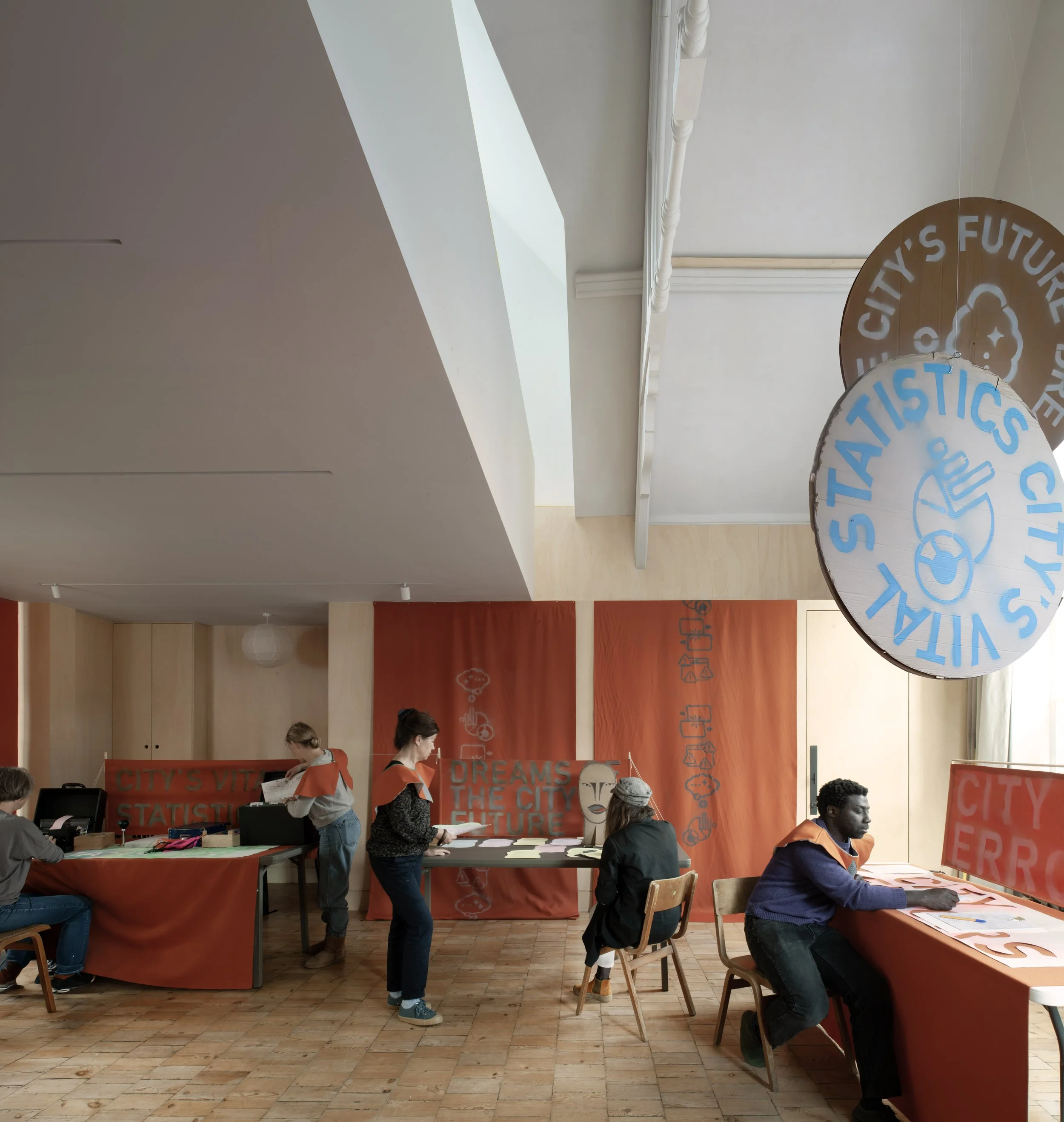 People attending a workshop or event in a room with orange banners and tables, engaging in activities such as reading, writing, and discussing. The room has wooden floors and a high ceiling with decorative moldings.