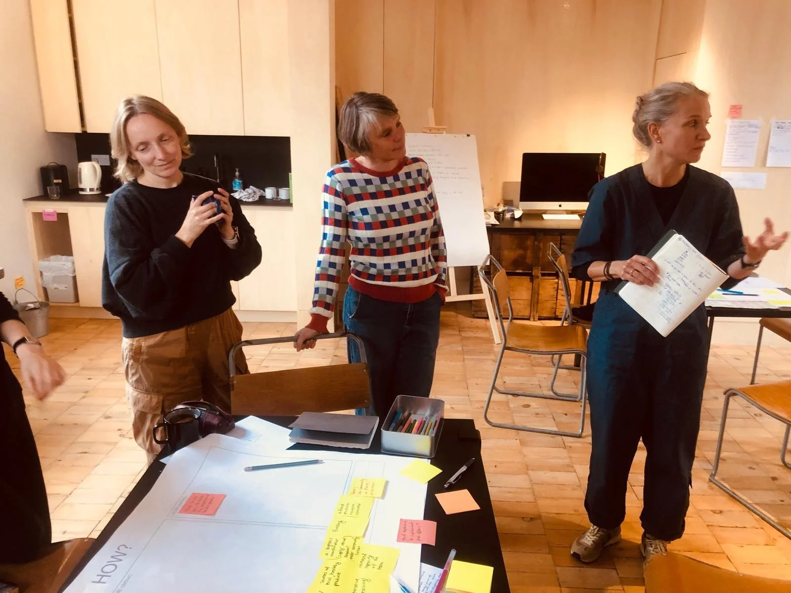 Three women standing in a room, engaging in a discussion or workshop, with chairs, a table with sticky notes and writing supplies, and a computer in the background.