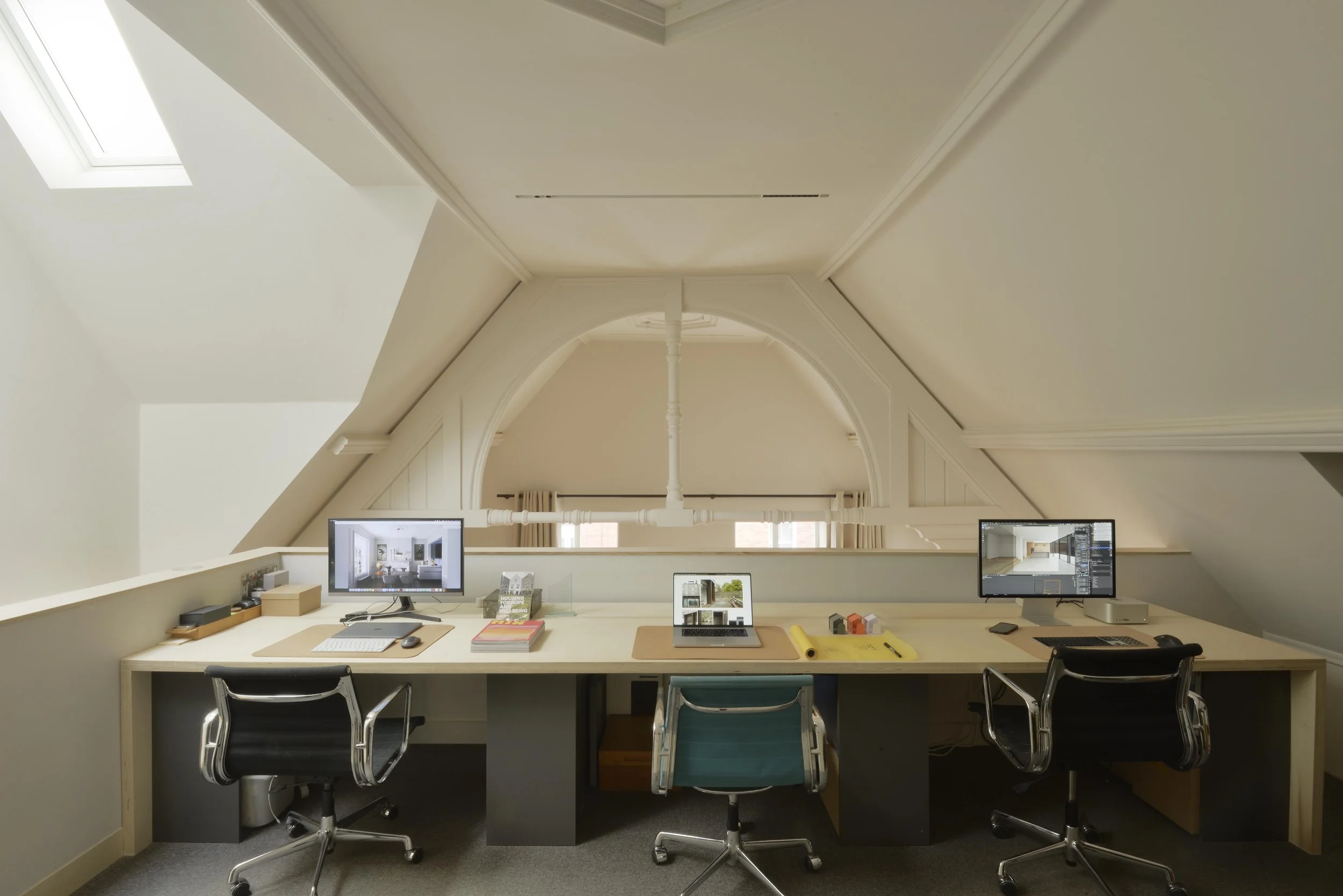 A home office workspace with three computer monitors on a long desk under an attic ceiling with skylights.