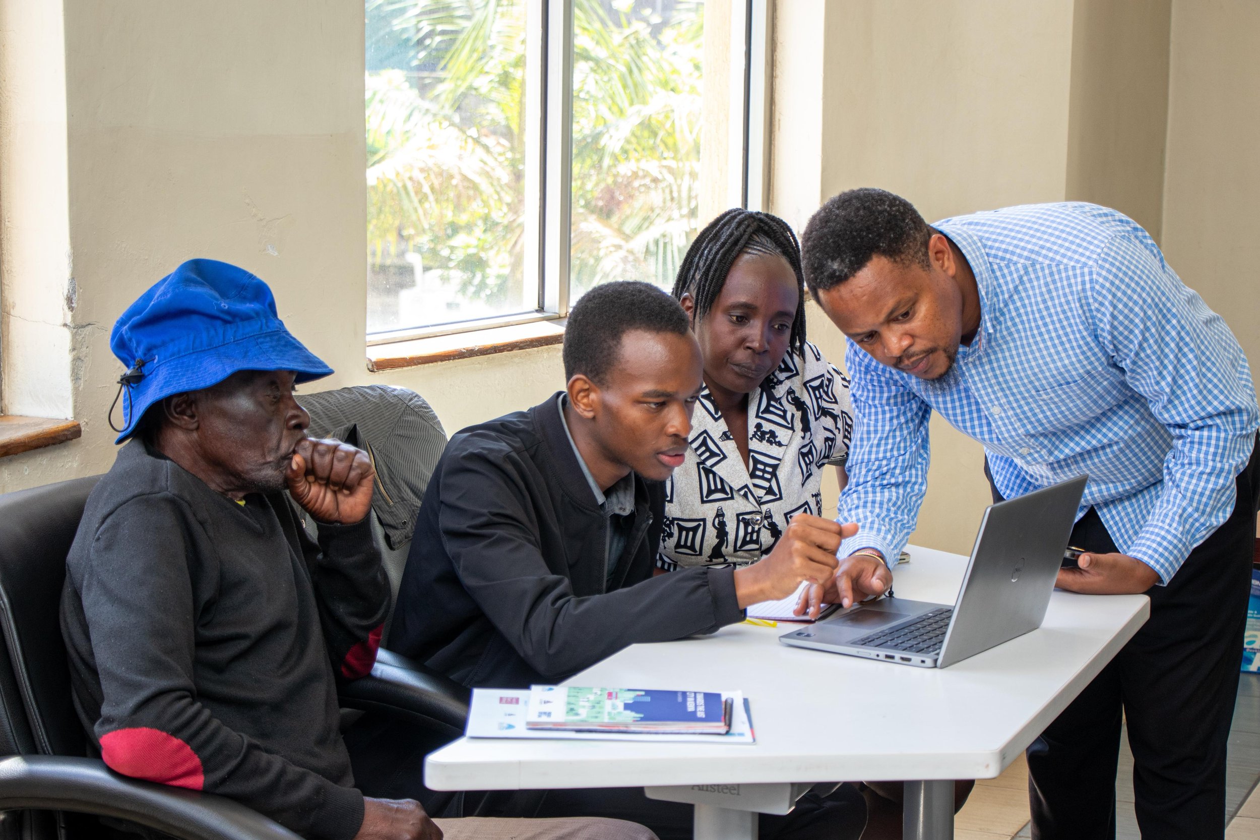 Four people working together around a laptop on a white table in a well-lit room with window blinds. One person is pointing at the laptop screen while others observe.