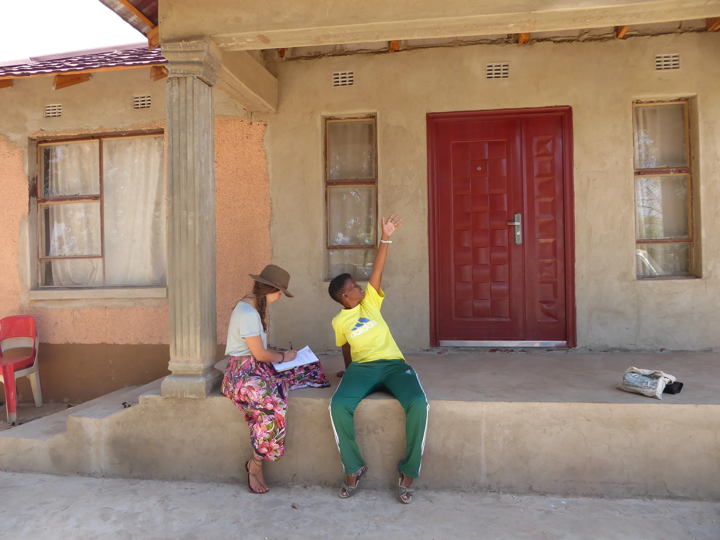 A woman is sitting on a step with a notebook, and a young boy in a yellow shirt is sitting on the same step, raising his hand. They are outside a house with a red door and two windows. A red chair and a bag are nearby.