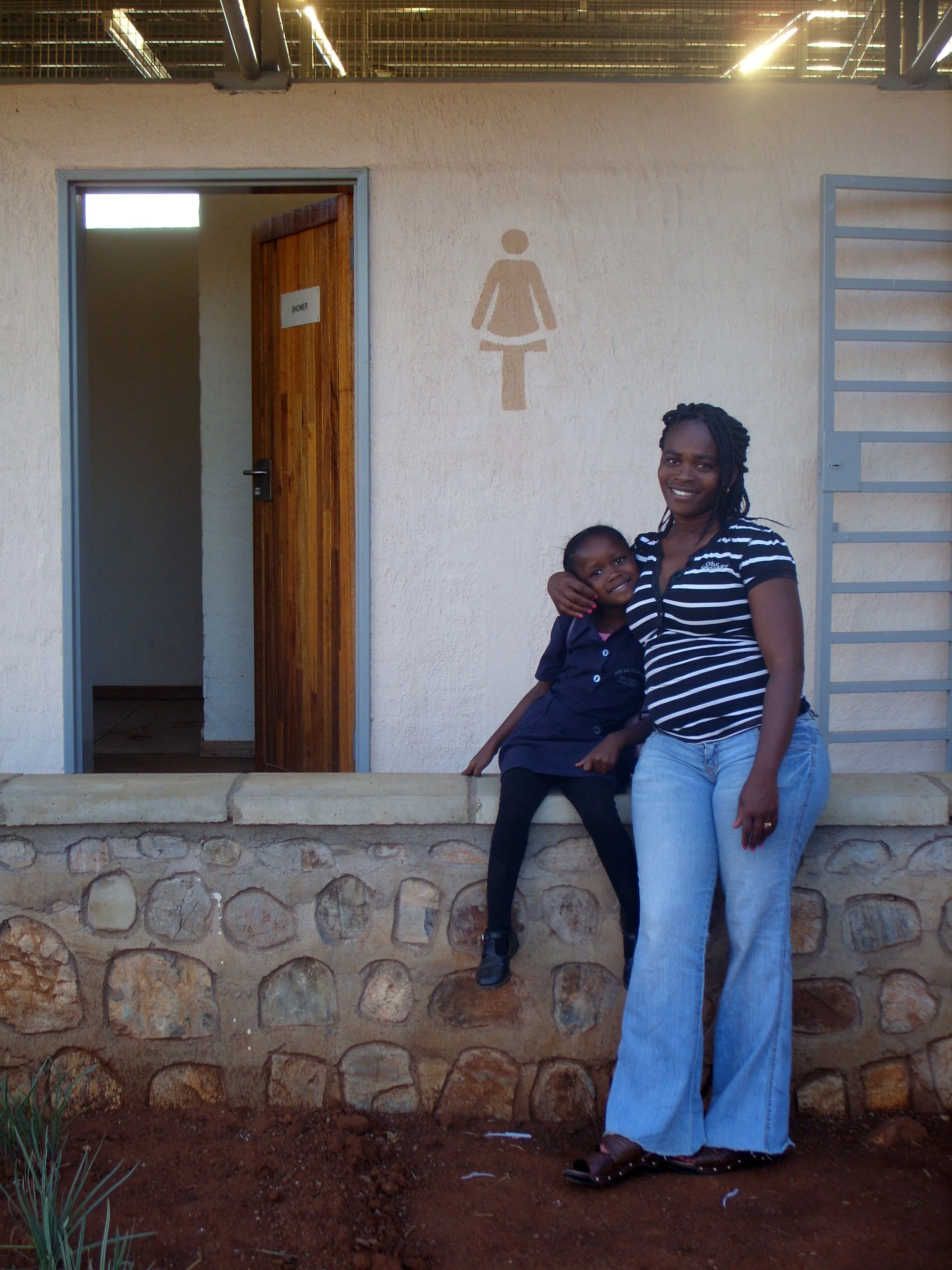 A woman and a young girl smiling and hugging in front of a building with a women's restroom sign.