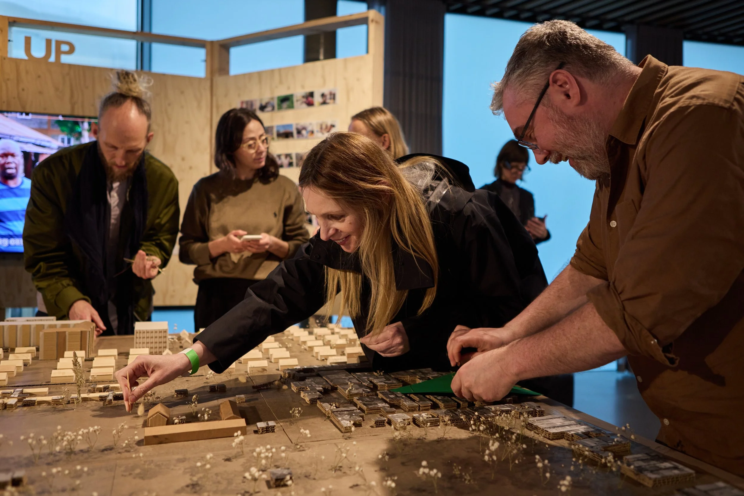 Group of people gathered around a large architectural model or urban planning display, examining and discussing it.