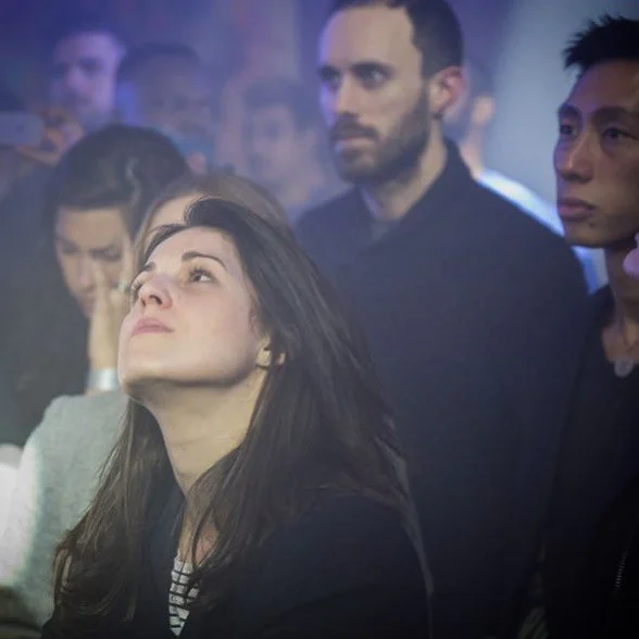 A group of diverse people at a concert or event, with some looking serious and others enjoying the moment, under dim lighting.