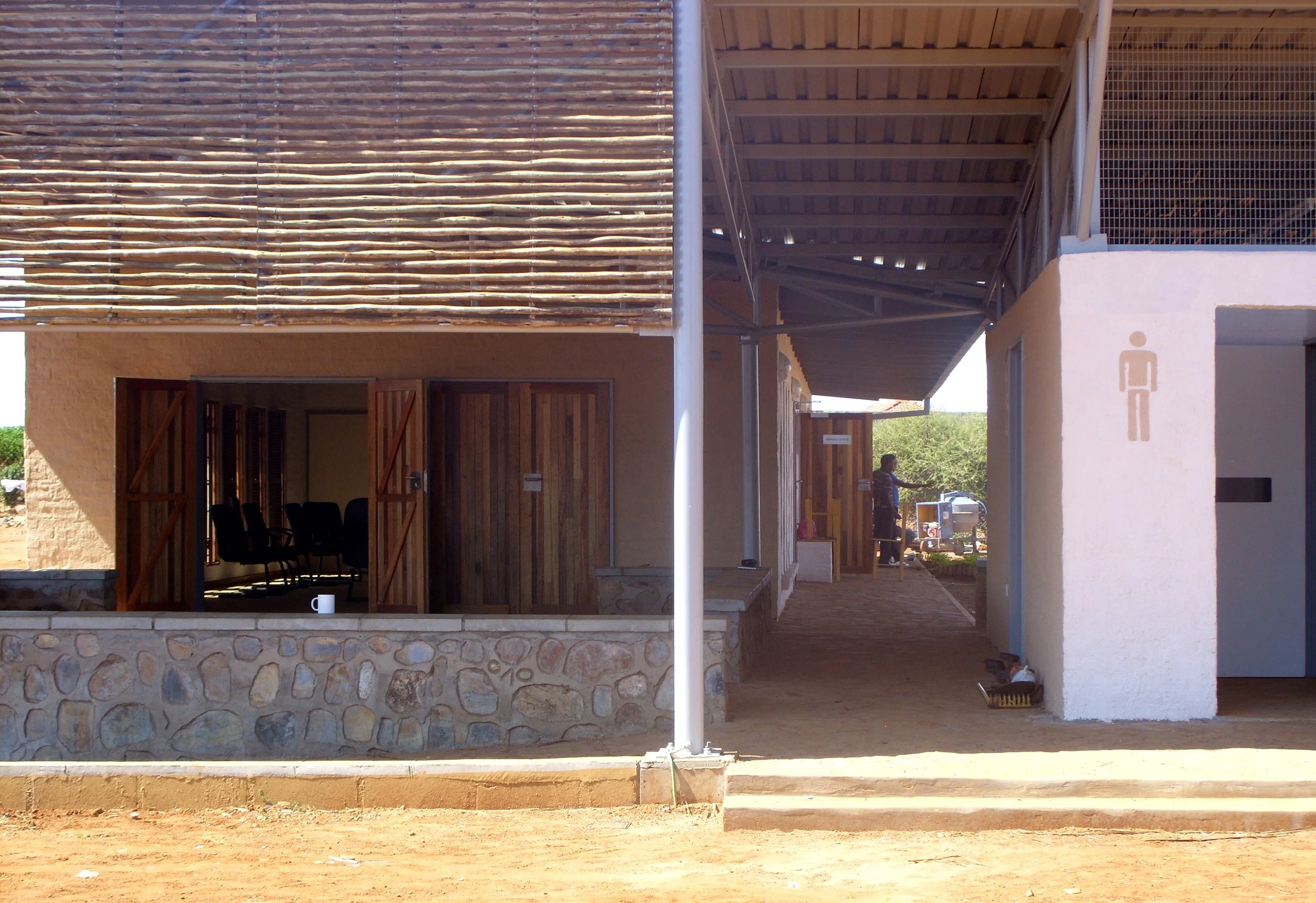 View of a building with wooden and stone exterior, open door revealing chairs, with a man standing outside near a food stand, and a restroom sign on a white wall.