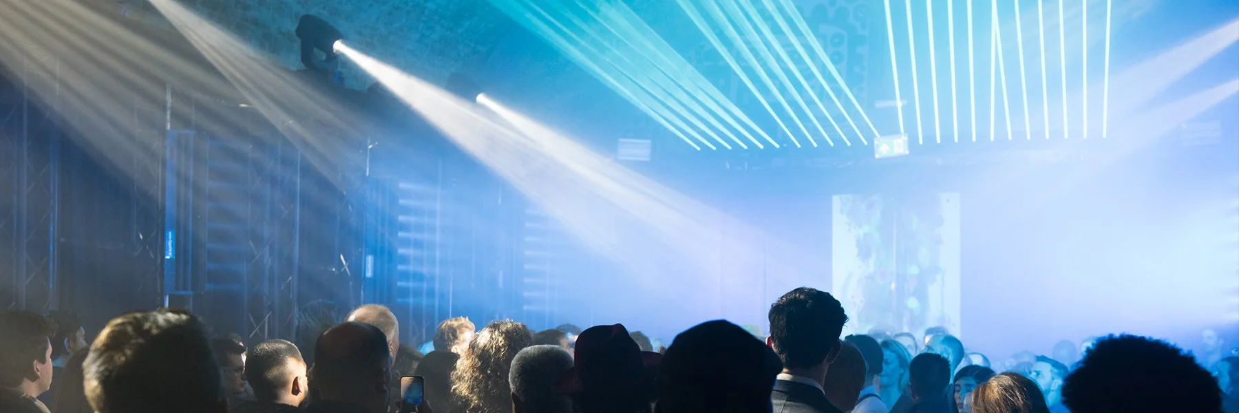 Audience sitting in a dark room watching a presentation or performance with blue lighting and stage lights.