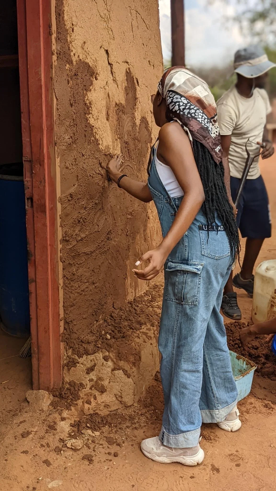 A woman with long dreadlocks, wearing a headscarf, white tank top, and overalls, is applying mud or clay to a wall outdoors. A man in a hat and casual clothes stands nearby, holding a tool, with a container placed on the ground.