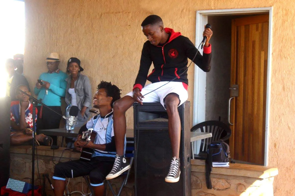 A young man playing a guitar while sitting on the lap of another young man, who is also holding a microphone, during a gathering with several people watching and taking pictures.