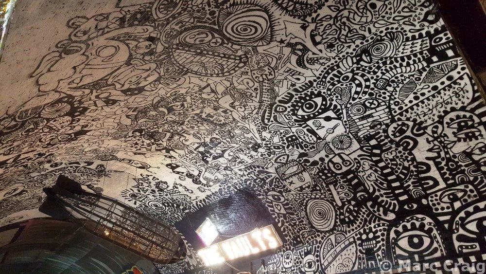 Black and white mural with intricate, abstract, and whimsical doodle-style illustrations covering a wall and ceiling, including eyes, faces, animals, and various abstract shapes.