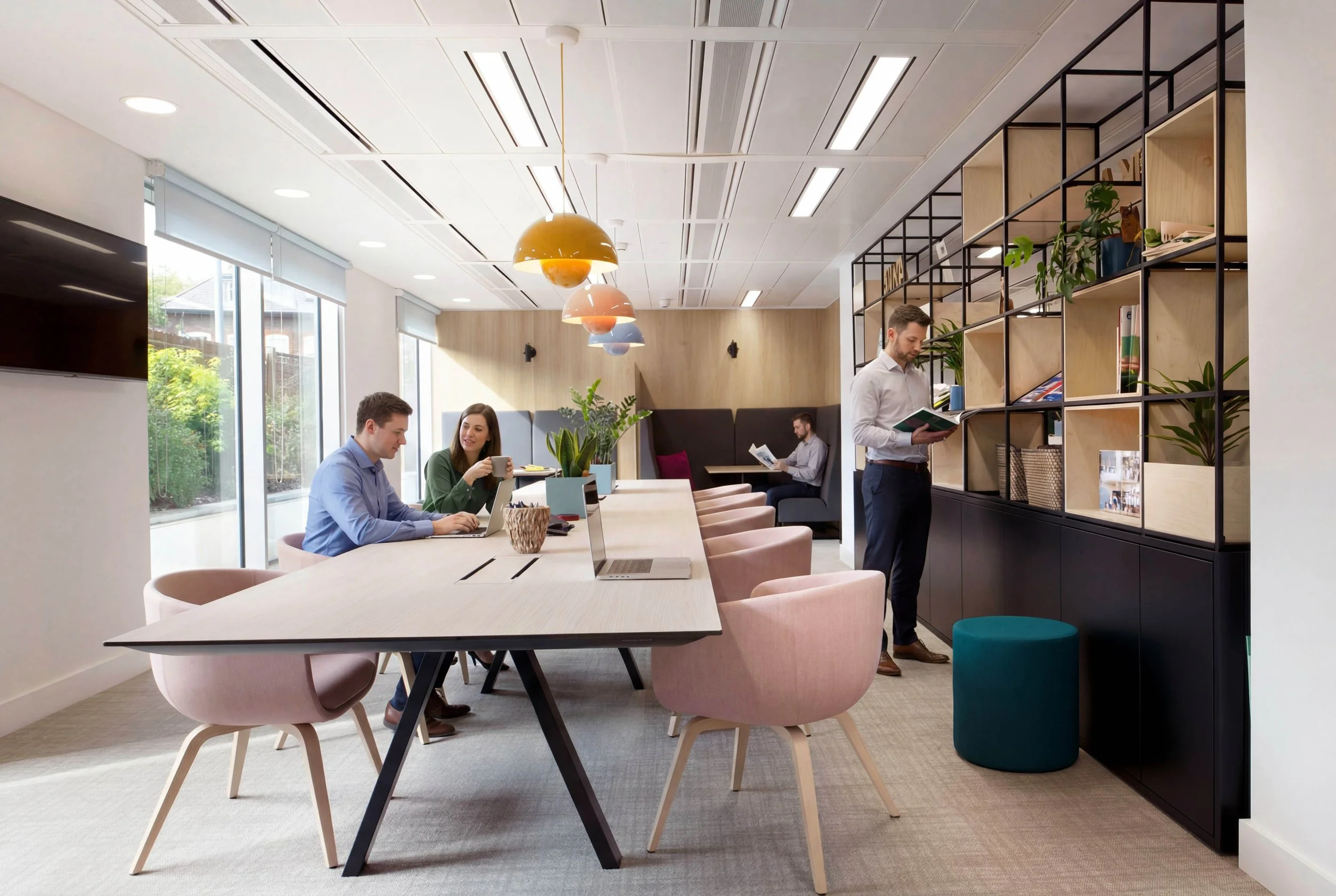 Modern coworking space with people working, large windows, and a black shelving unit with plants and books.