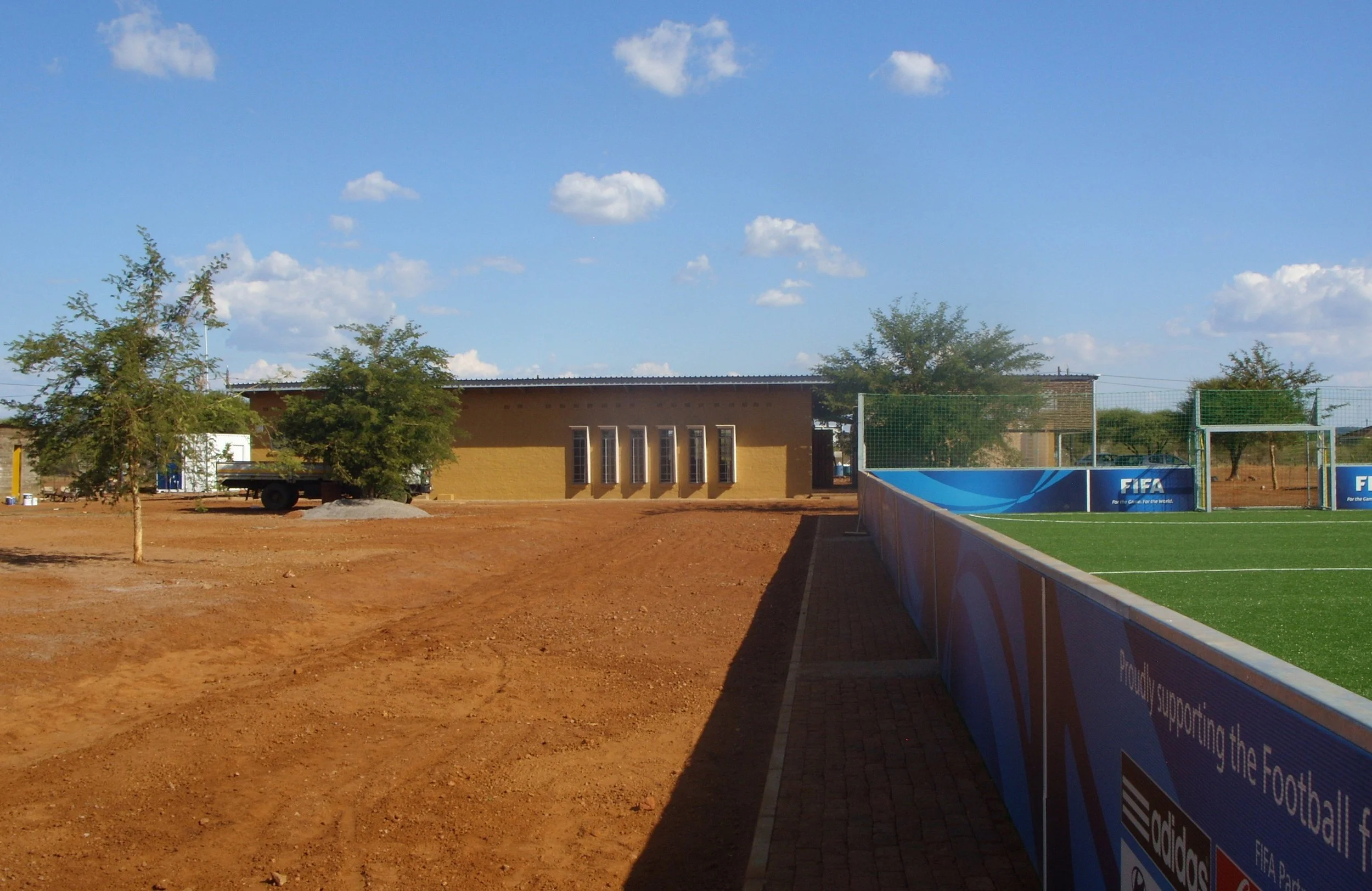 A soccer field with green artificial turf, surrounded by a blue advertising barrier, next to a building with rectangular windows, and trees, under a partly cloudy sky.