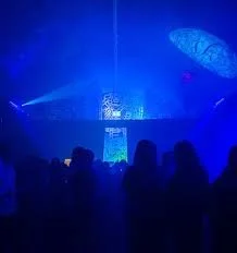 A nightclub or concert venue with blue lighting, silhouettes of people watching a performance on stage, with some geometric decorative elements overhead.