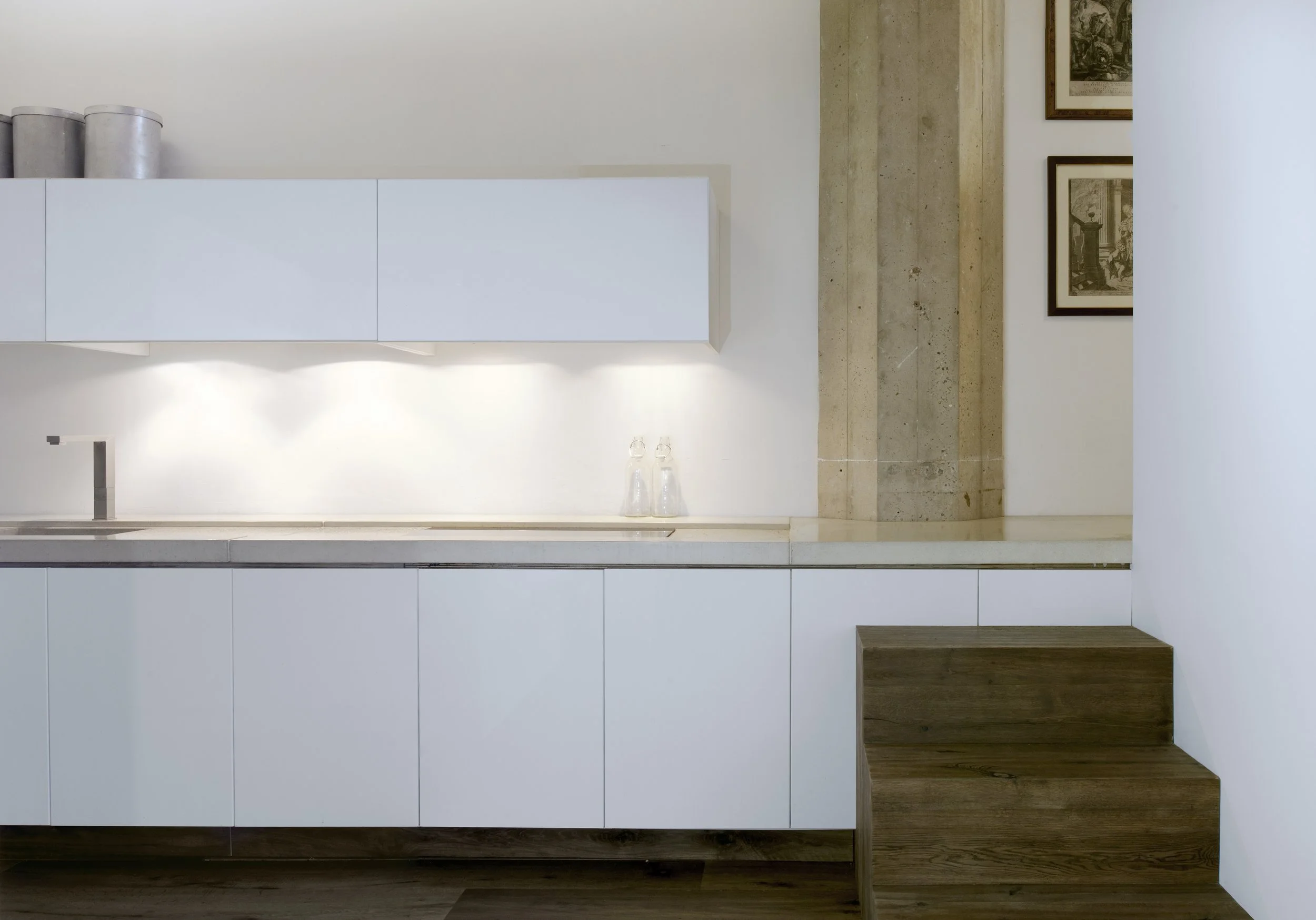 Minimalist kitchen with white cabinets, a wooden step, and framed black-and-white artwork on the wall.