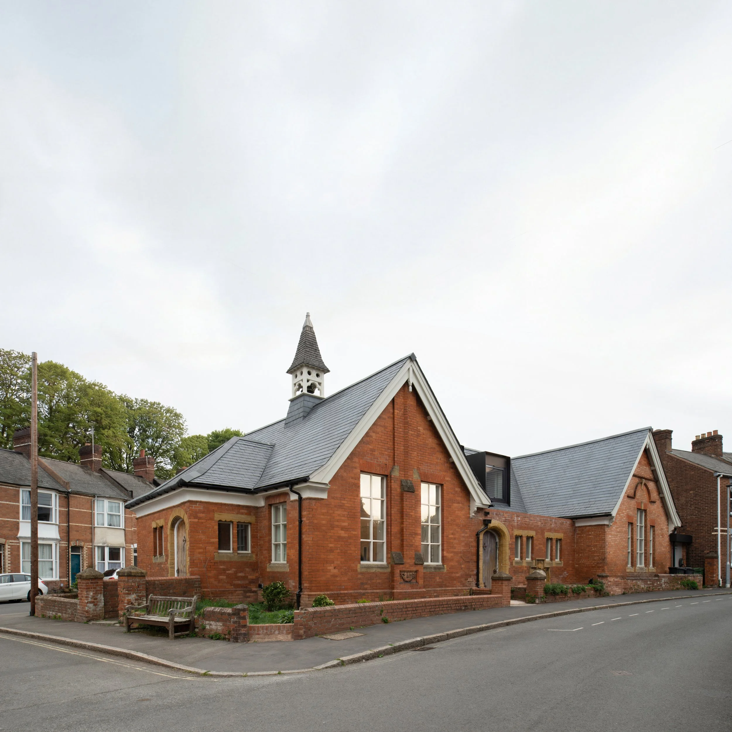 A brick church with a steep peaked roof and a small tower on top, situated on a corner with a tree-lined background and a sidewalk in front.