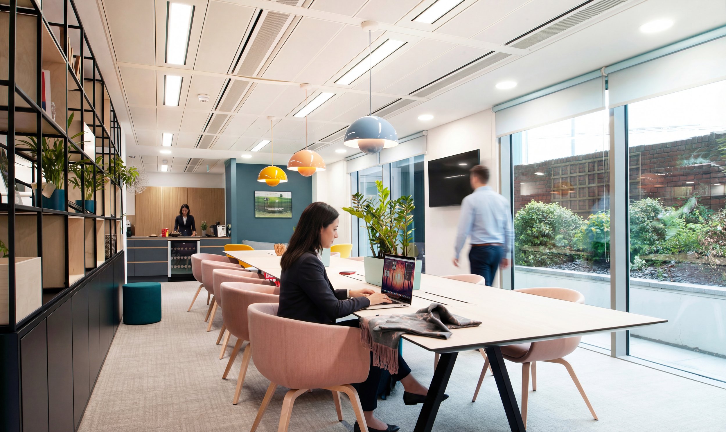 Office break room with large window, pink chairs at a long white table, a woman working on a laptop, a man walking, a woman in the background near a counter, colorful pendant lights, TV on the wall, plants, and shelving with decorative items.